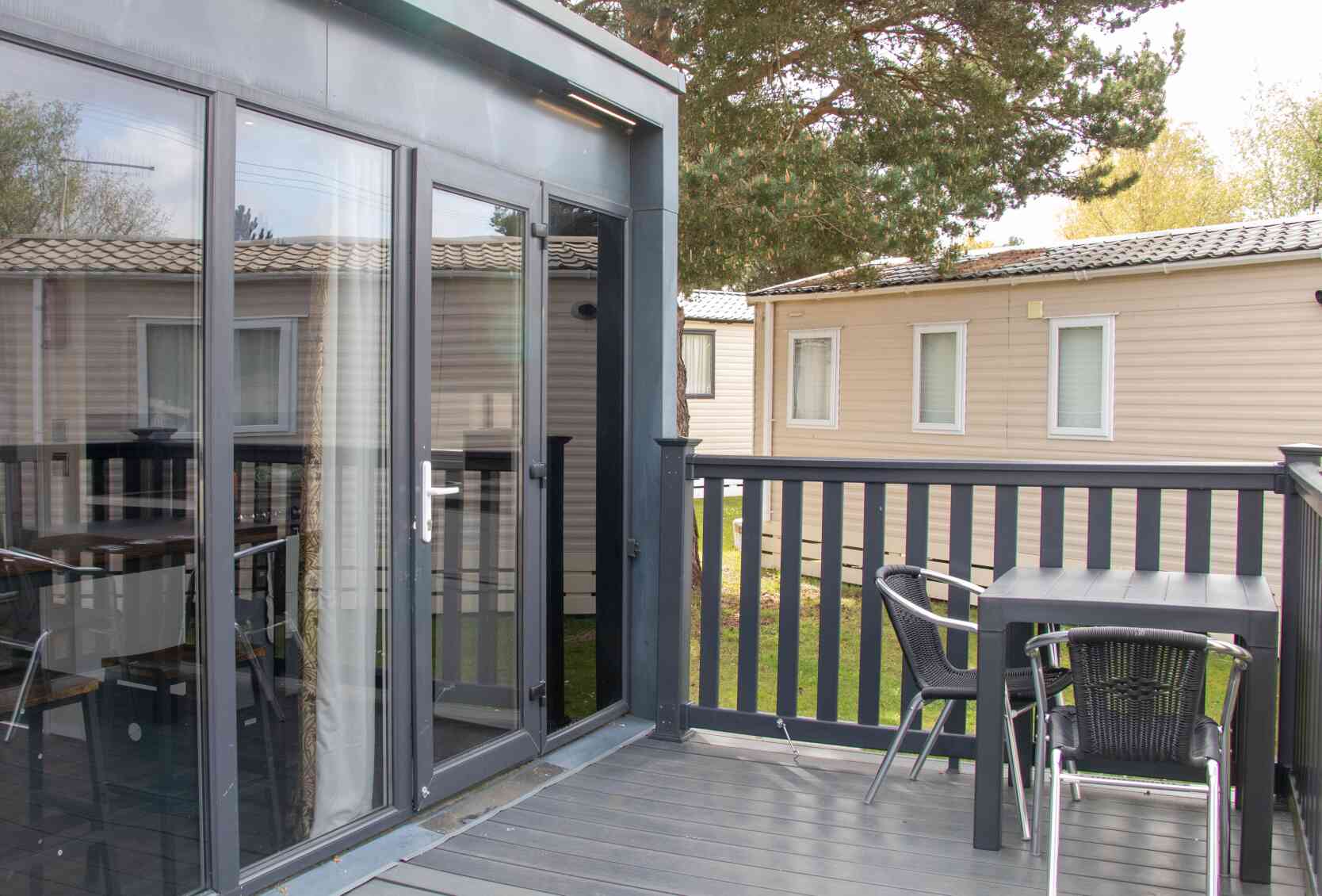 A modern deck area featuring a table and chairs, with large glass doors leading to an interior space. In the background, there are several beige cabins and green trees.