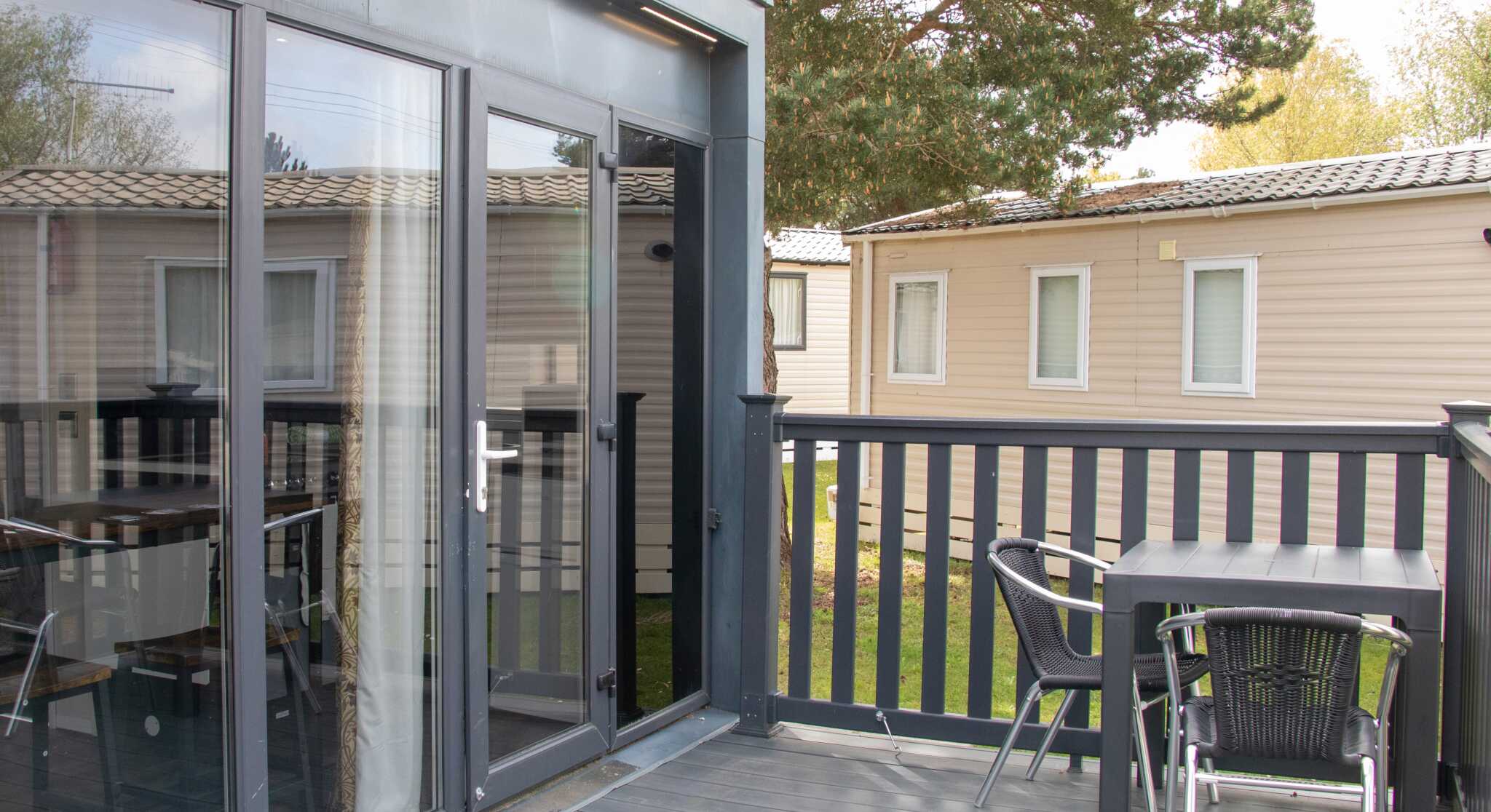 A modern deck area featuring a table and chairs, with large glass doors leading to an interior space. In the background, there are several beige cabins and green trees.