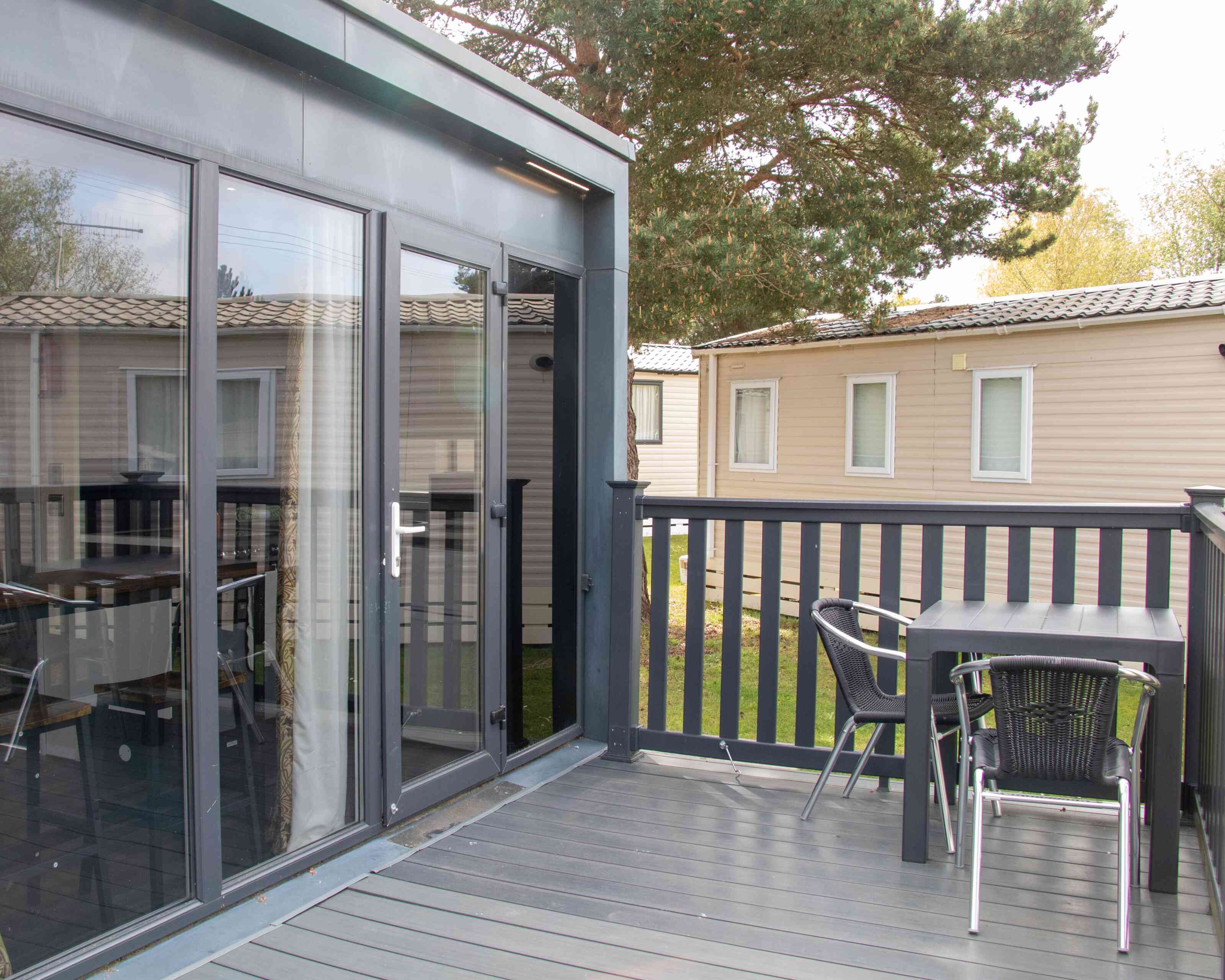 A modern deck area featuring a table and chairs, with large glass doors leading to an interior space. In the background, there are several beige cabins and green trees.