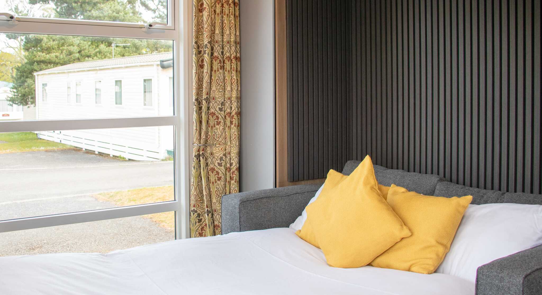 A cozy bedroom features a neatly made bed with white linens and two yellow pillows. A large window lets in natural light and offers a view of a mobile home park outside. The wall behind the bed has a vertical panel design, and there are elegant curtains.