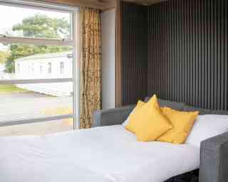 A cozy bedroom features a neatly made bed with white linens and two yellow pillows. A large window lets in natural light and offers a view of a mobile home park outside. The wall behind the bed has a vertical panel design, and there are elegant curtains.