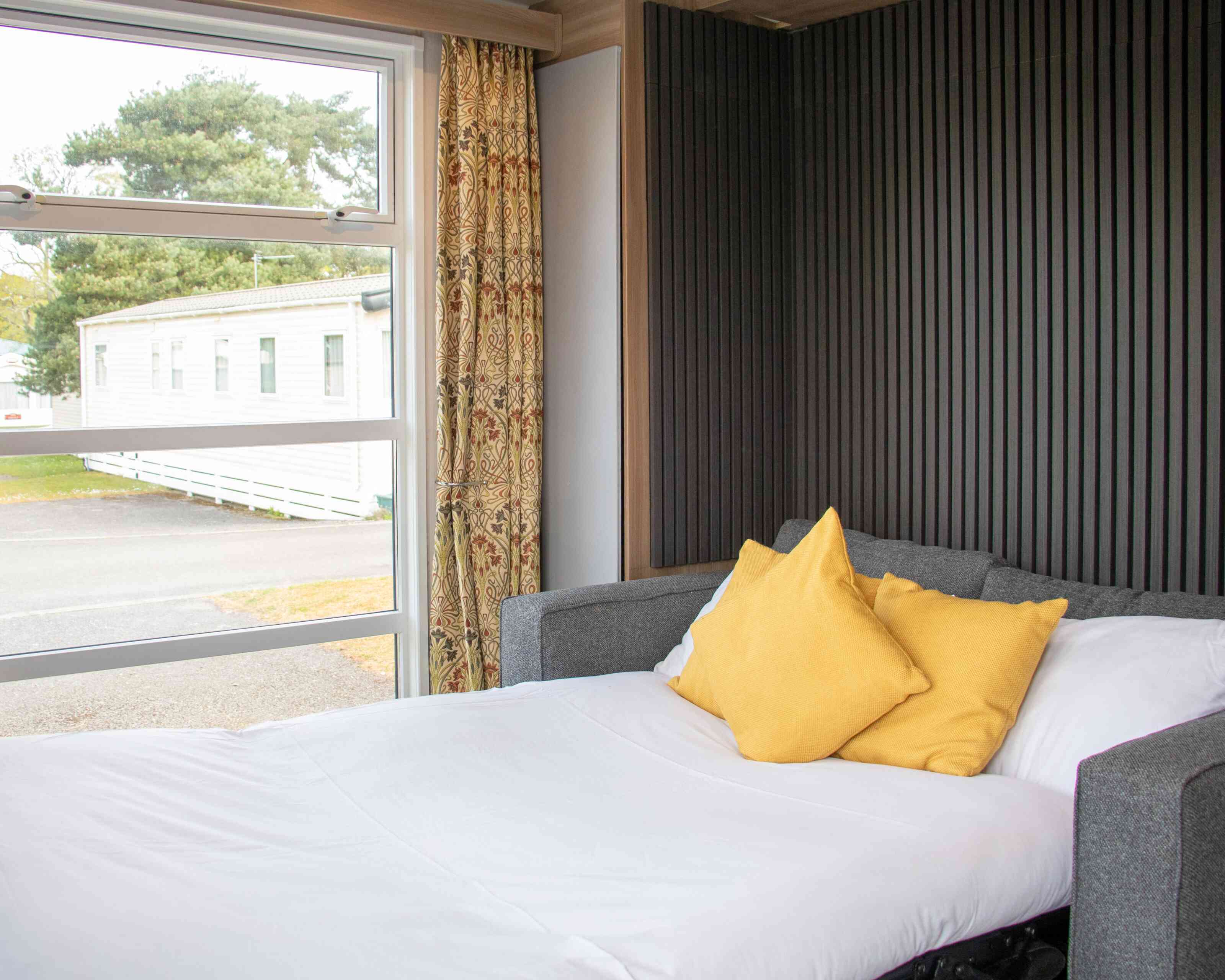 A cozy bedroom features a neatly made bed with white linens and two yellow pillows. A large window lets in natural light and offers a view of a mobile home park outside. The wall behind the bed has a vertical panel design, and there are elegant curtains.