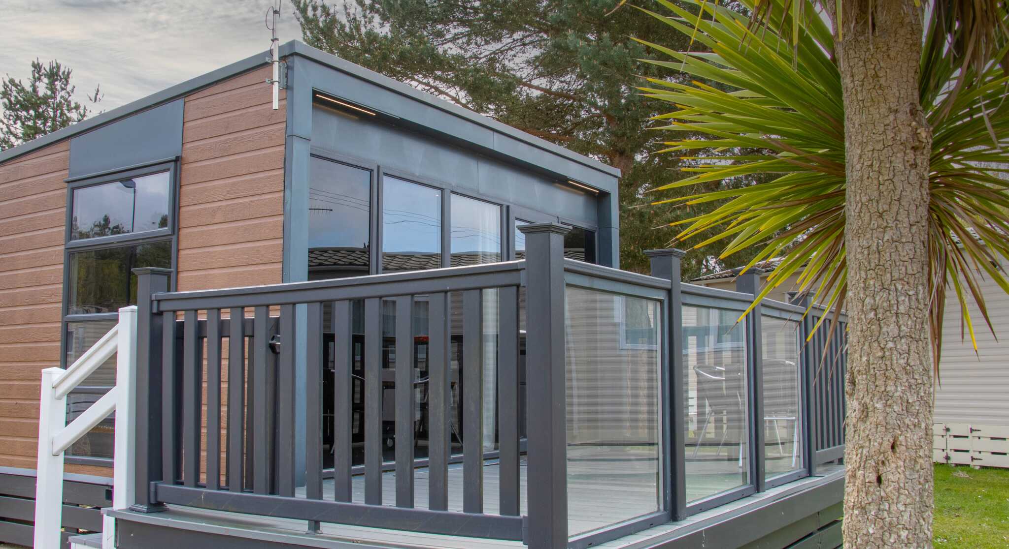 A modern cabin with large windows and a gray deck, featuring a glass railing. Lush green palm-like plants are in the foreground, and a cloudy sky is visible above.