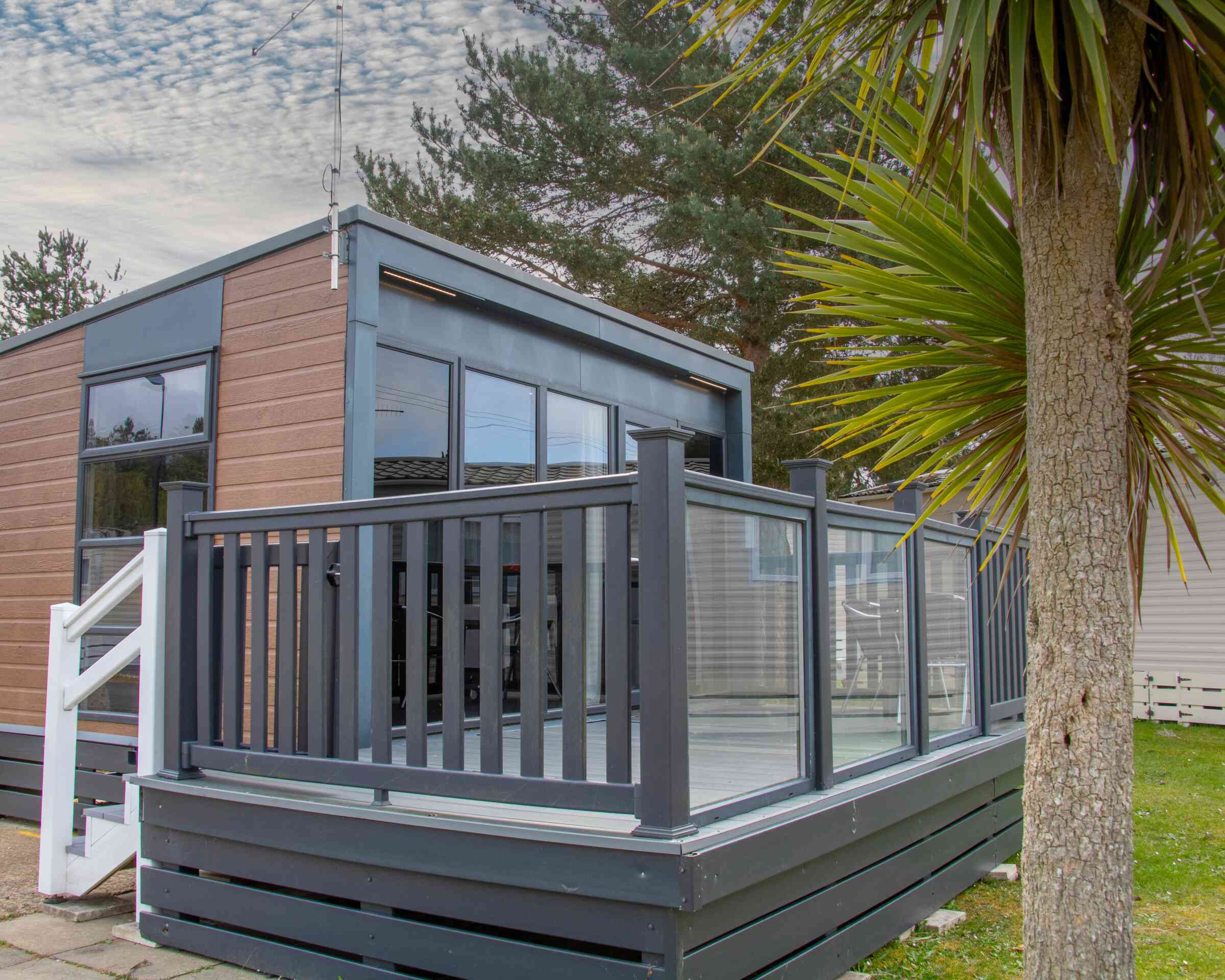 A modern cabin with large windows and a gray deck, featuring a glass railing. Lush green palm-like plants are in the foreground, and a cloudy sky is visible above.