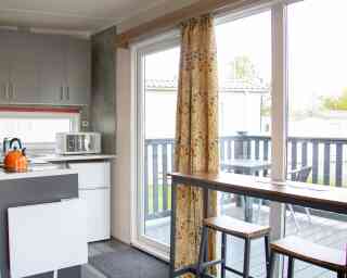 A modern kitchen area featuring gray cabinets, a microwave, and a small refrigerator. There's a sliding glass door with colorful curtains leading to a deck, and a wooden dining table with two stools. Outdoor seating is visible on the deck.