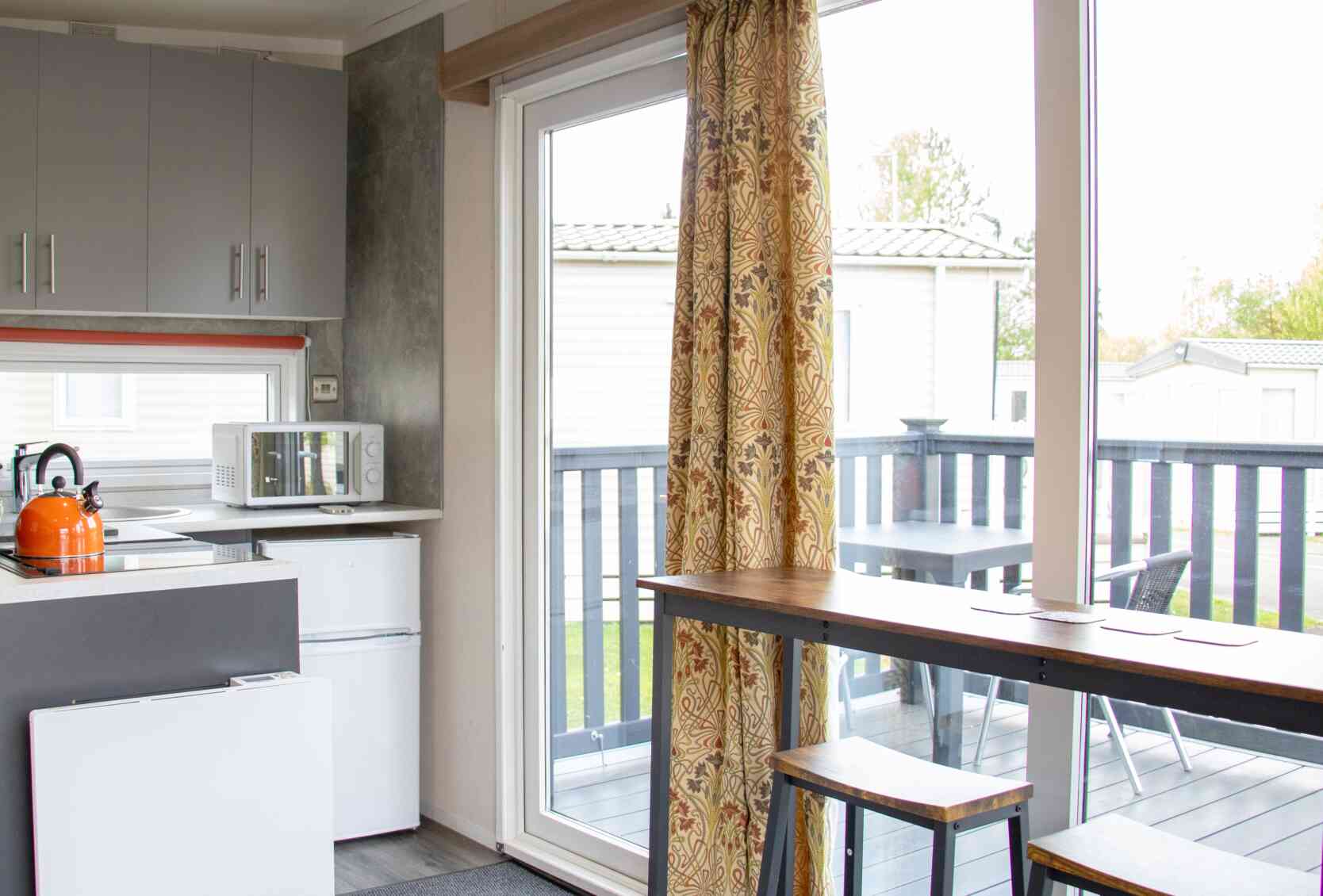 A modern kitchen area featuring gray cabinets, a microwave, and a small refrigerator. There's a sliding glass door with colorful curtains leading to a deck, and a wooden dining table with two stools. Outdoor seating is visible on the deck.