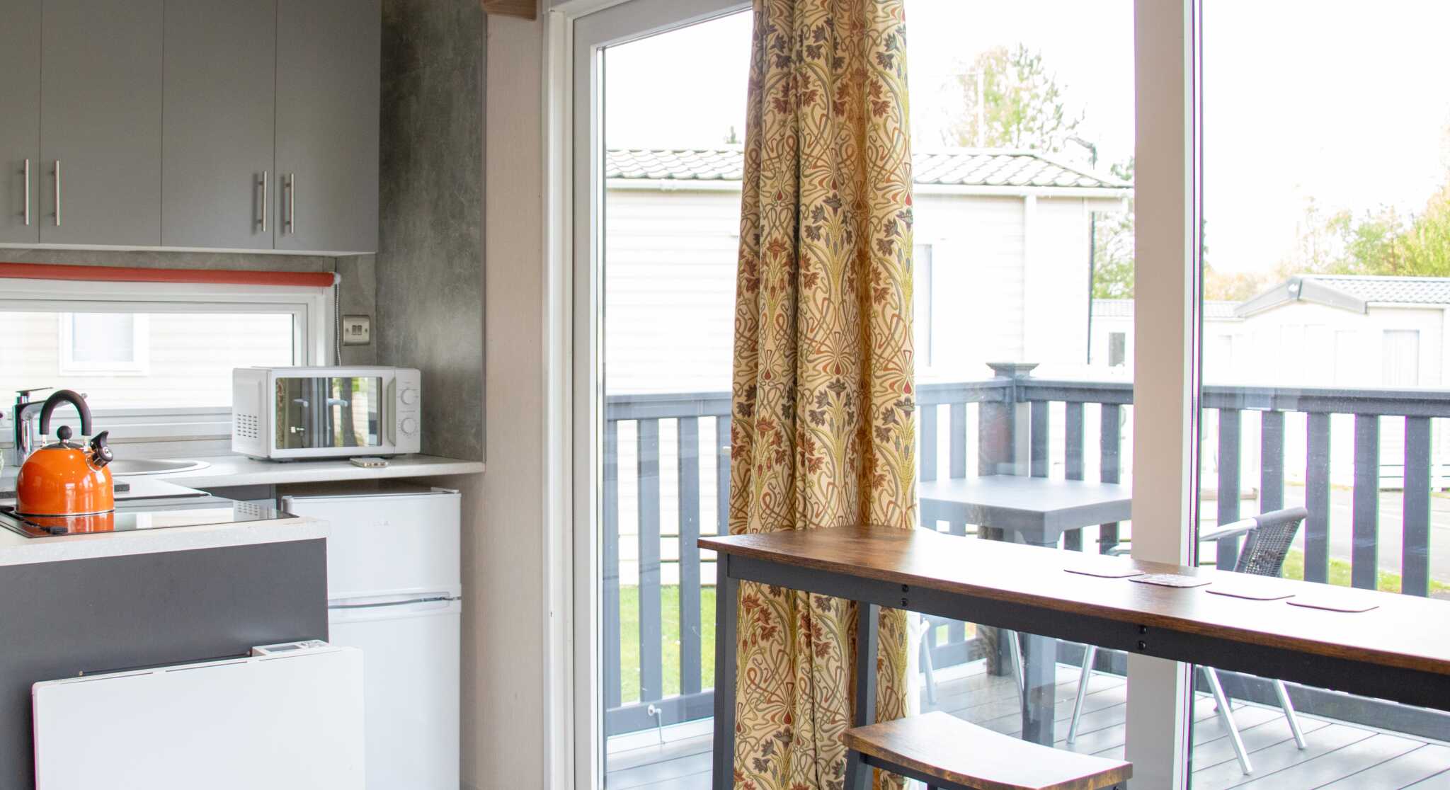 A modern kitchen area featuring gray cabinets, a microwave, and a small refrigerator. There's a sliding glass door with colorful curtains leading to a deck, and a wooden dining table with two stools. Outdoor seating is visible on the deck.
