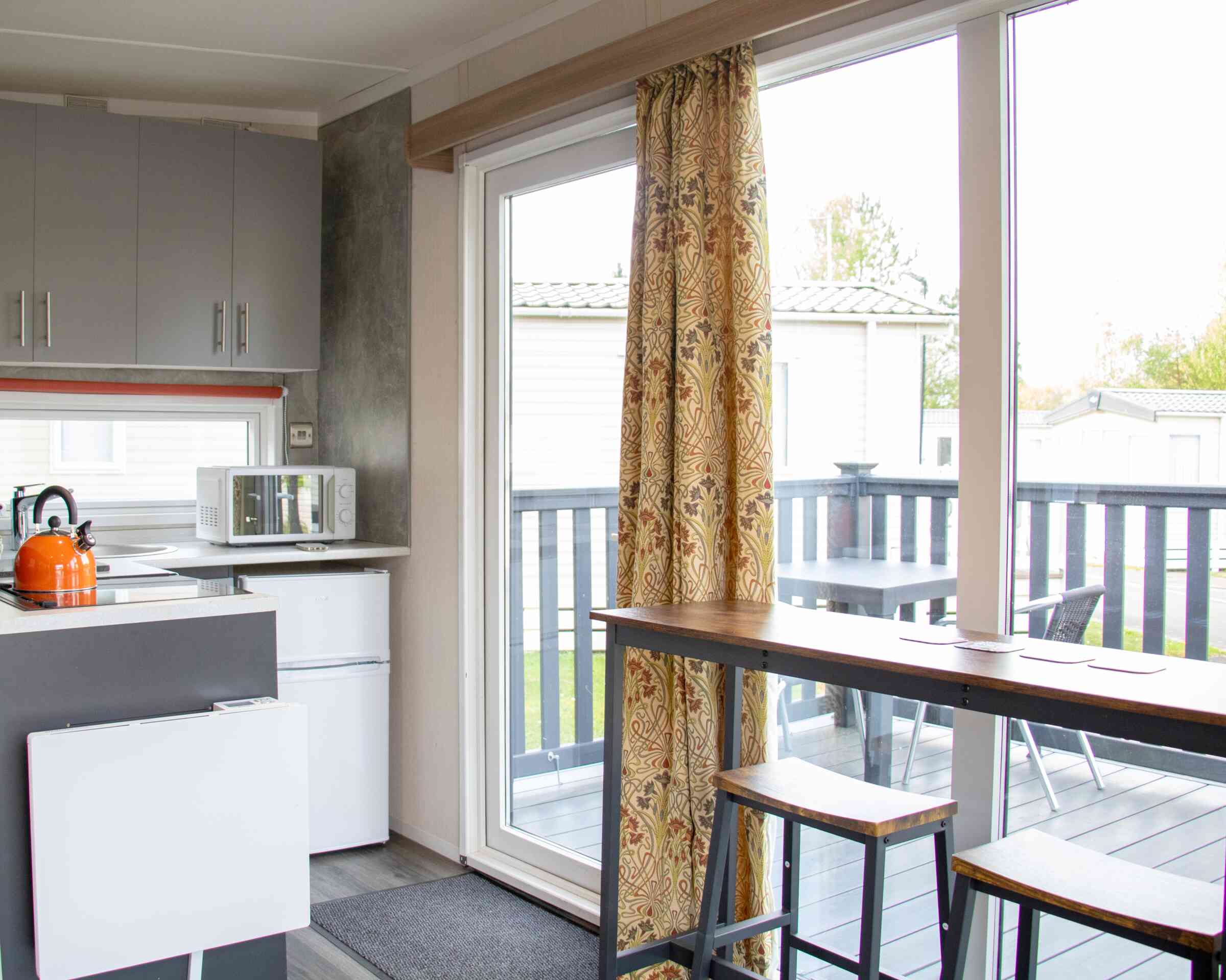 A modern kitchen area featuring gray cabinets, a microwave, and a small refrigerator. There's a sliding glass door with colorful curtains leading to a deck, and a wooden dining table with two stools. Outdoor seating is visible on the deck.