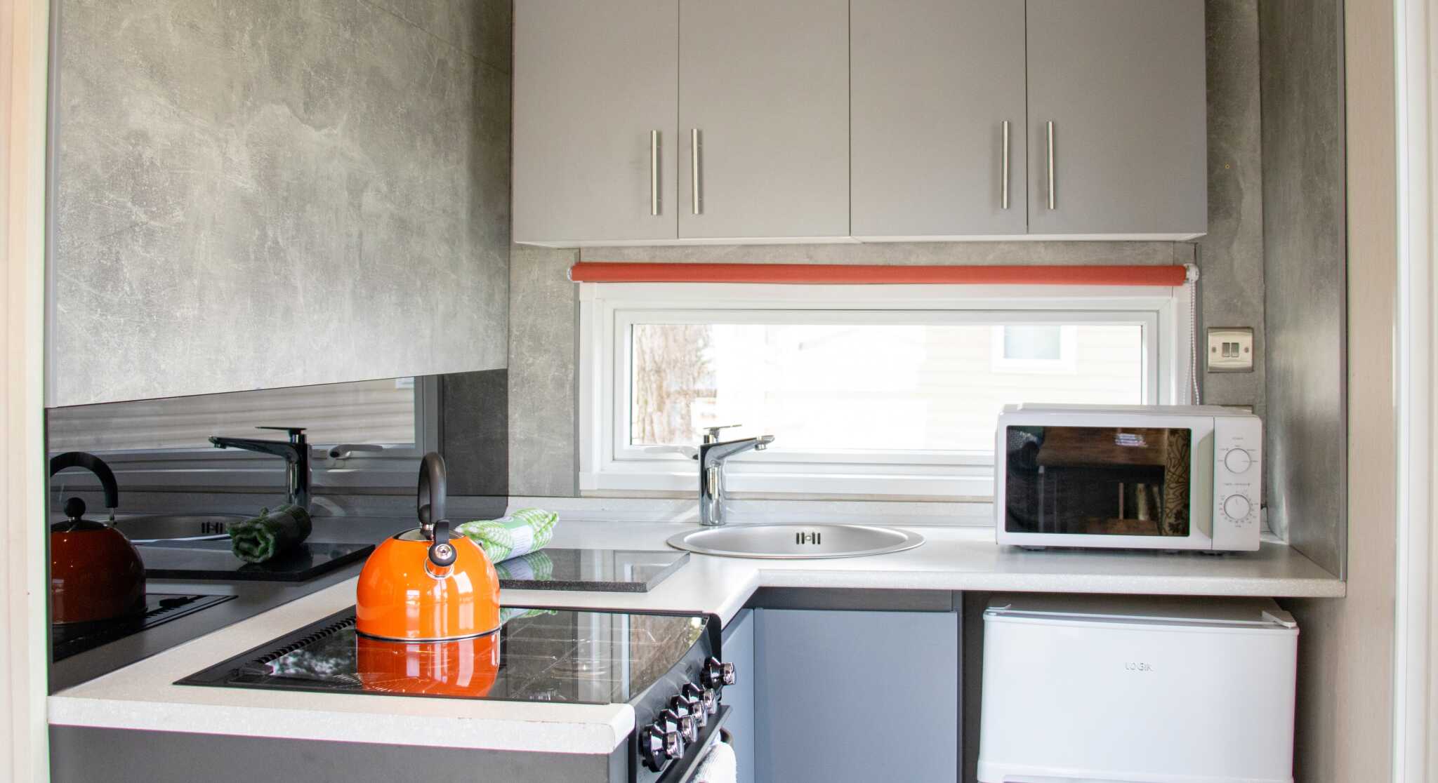 A modern kitchen featuring gray cabinets, a sleek countertop, an orange kettle on a stove, a sink beneath a window, and a white refrigerator.