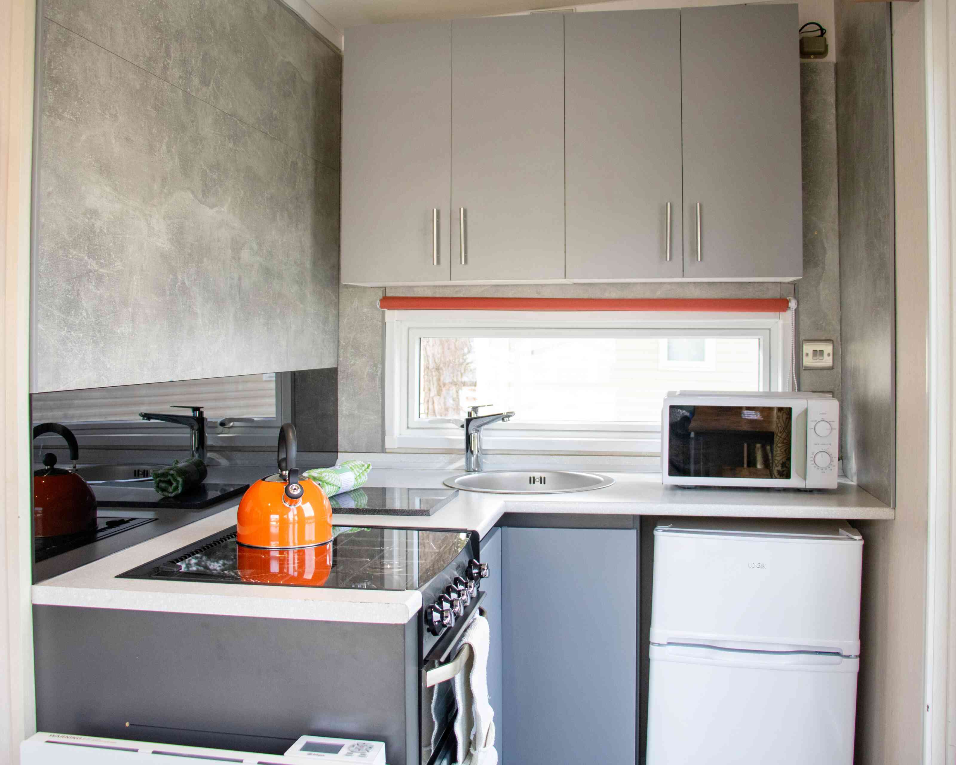 A modern kitchen featuring gray cabinets, a sleek countertop, an orange kettle on a stove, a sink beneath a window, and a white refrigerator.