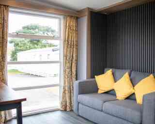 A cozy interior featuring a gray sofa with yellow cushions, large window with sheer curtains, and a view of the outdoors through the window. The walls have a modern wooden and textured design.