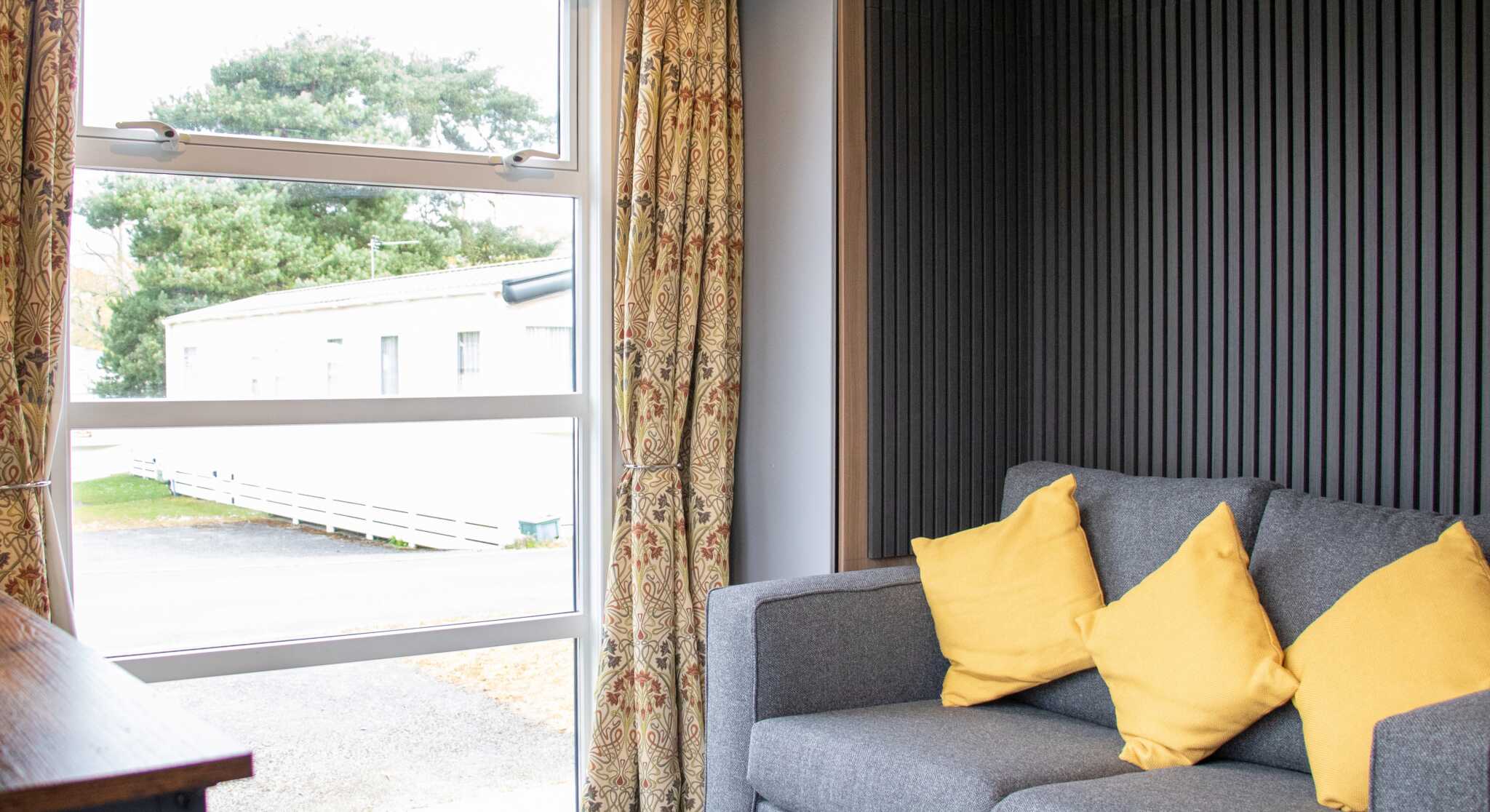 A cozy interior featuring a gray sofa with yellow cushions, large window with sheer curtains, and a view of the outdoors through the window. The walls have a modern wooden and textured design.