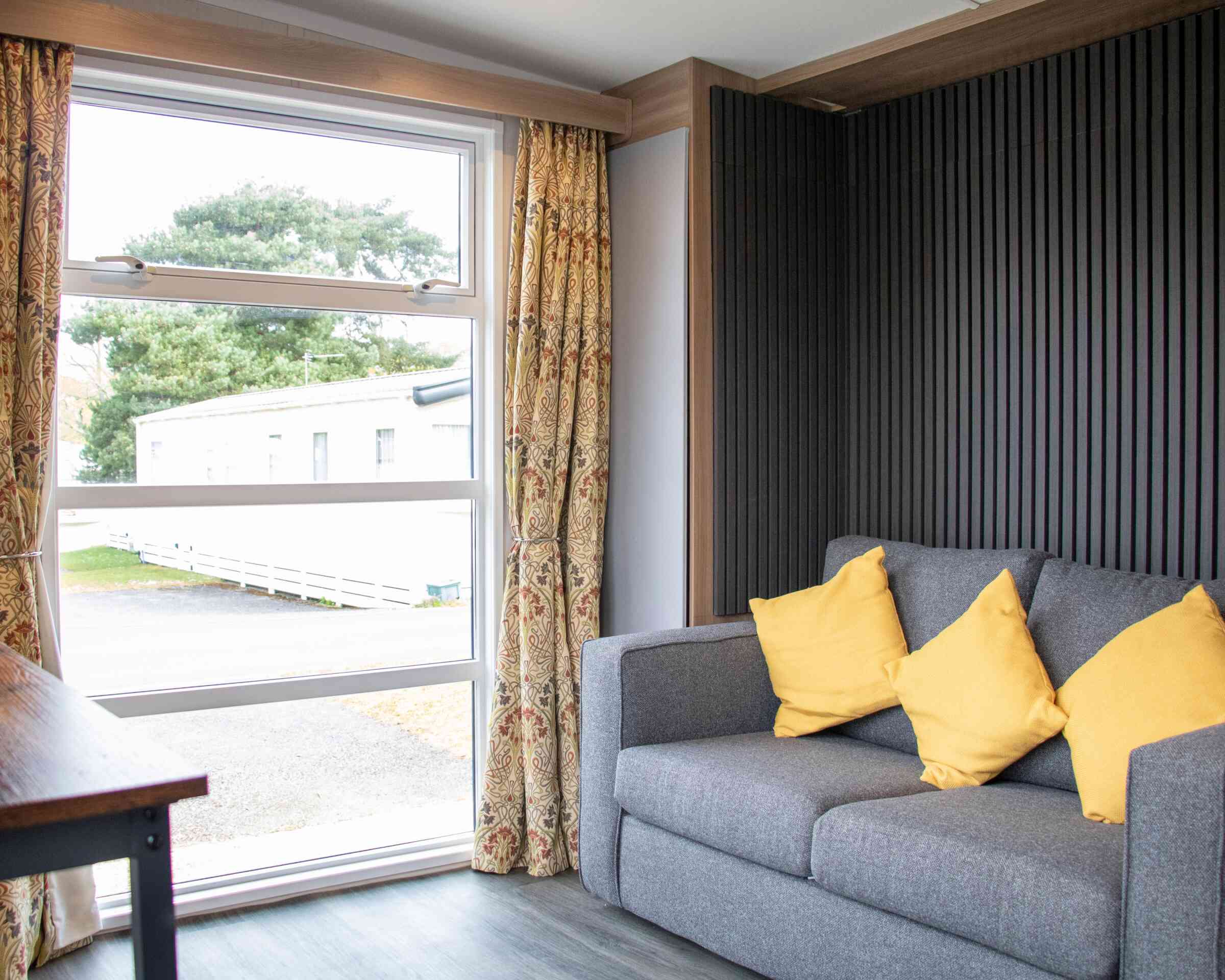 A cozy interior featuring a gray sofa with yellow cushions, large window with sheer curtains, and a view of the outdoors through the window. The walls have a modern wooden and textured design.
