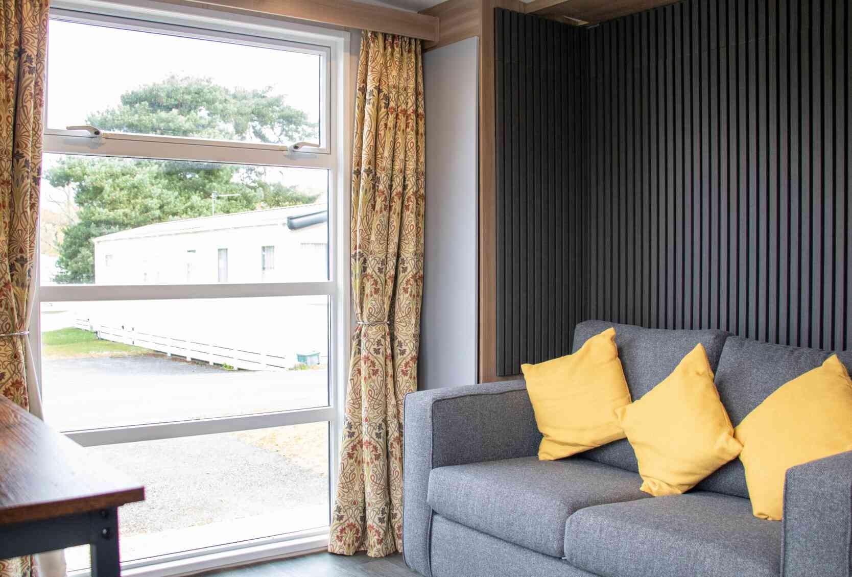 A cozy interior featuring a gray sofa with yellow cushions, large window with sheer curtains, and a view of the outdoors through the window. The walls have a modern wooden and textured design.