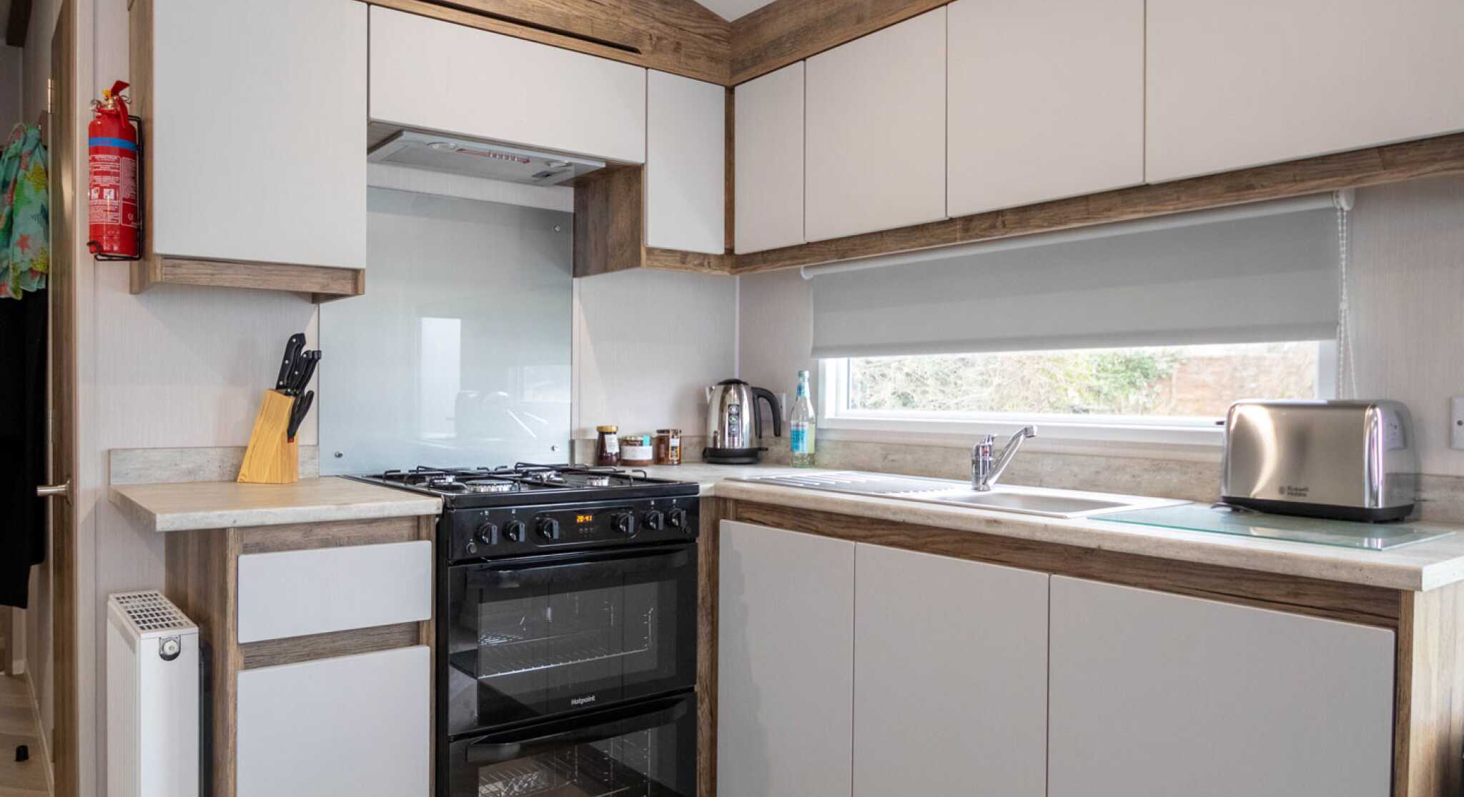 Modern kitchen with a combination of light gray and wooden cabinetry. Features a black oven and stovetop, stainless steel kettle, and a silver toaster. A large window provides natural light, and there's a small radiator on the wall.