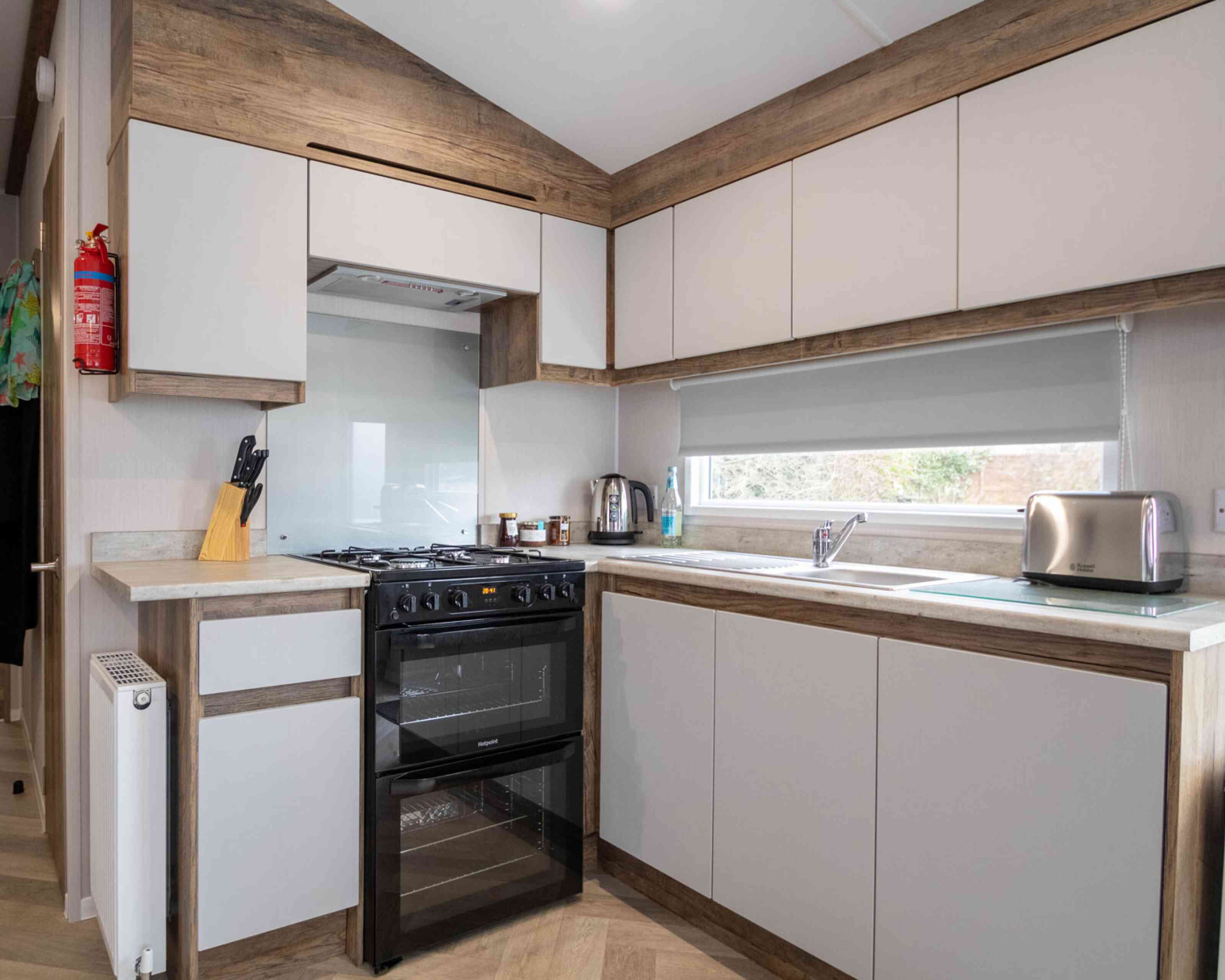 Modern kitchen with a combination of light gray and wooden cabinetry. Features a black oven and stovetop, stainless steel kettle, and a silver toaster. A large window provides natural light, and there's a small radiator on the wall.