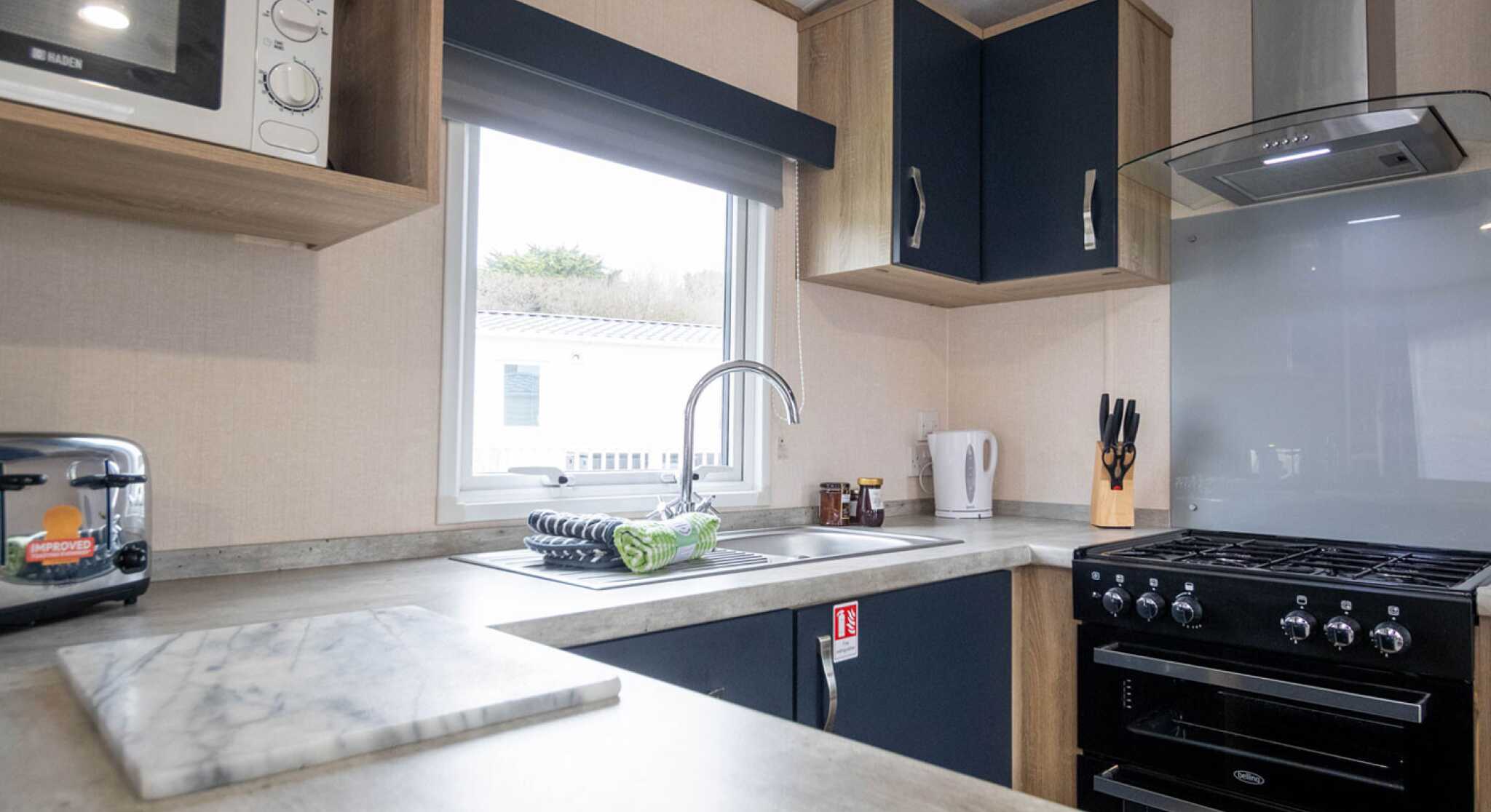 A modern kitchen with wooden cabinets and a grey countertop. The space features a sink with a stylish faucet, a gas stove, a microwave, and a window with natural light. Kitchen utensils are neatly arranged near the stove, and there's a marble cutting board on the counter.