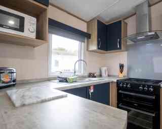 A modern kitchen with wooden cabinets and a grey countertop. The space features a sink with a stylish faucet, a gas stove, a microwave, and a window with natural light. Kitchen utensils are neatly arranged near the stove, and there's a marble cutting board on the counter.