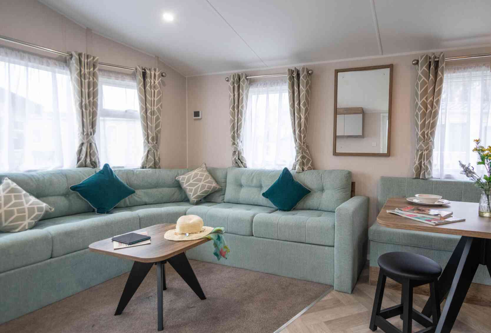 A cozy living area featuring a light teal sectional sofa with decorative pillows, a wooden coffee table with a book and a sunhat, and a dining table set for two. Sunlight streams through sheer curtains, illuminating the space.