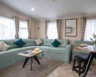 A cozy living area featuring a light teal sectional sofa with decorative pillows, a wooden coffee table with a book and a sunhat, and a dining table set for two. Sunlight streams through sheer curtains, illuminating the space.