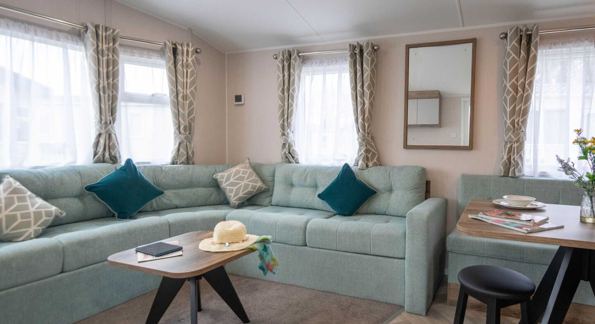 A cozy living area featuring a light teal sectional sofa with decorative pillows, a wooden coffee table with a book and a sunhat, and a dining table set for two. Sunlight streams through sheer curtains, illuminating the space.