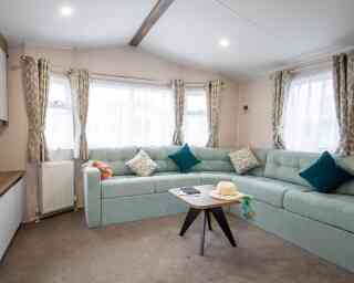 A cozy living room featuring a light blue sectional sofa with decorative pillows, large windows with sheer curtains, and a modern wooden coffee table. A sun hat and a notebook are placed on the table. The space has a warm and inviting atmosphere.