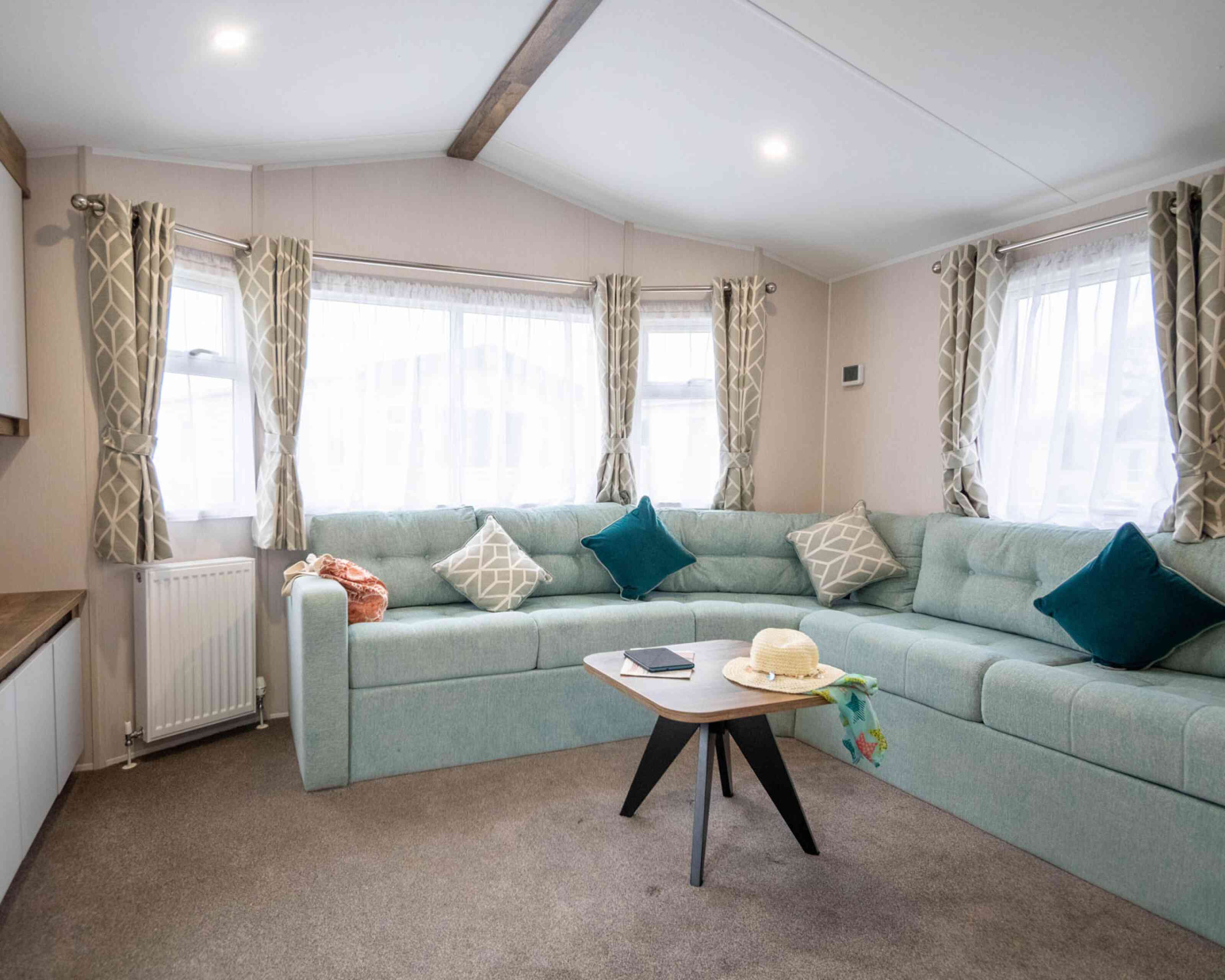 A cozy living room featuring a light blue sectional sofa with decorative pillows, large windows with sheer curtains, and a modern wooden coffee table. A sun hat and a notebook are placed on the table. The space has a warm and inviting atmosphere.