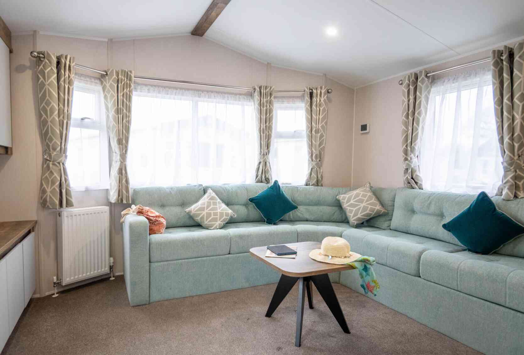 A cozy living room featuring a light blue sectional sofa with decorative pillows, large windows with sheer curtains, and a modern wooden coffee table. A sun hat and a notebook are placed on the table. The space has a warm and inviting atmosphere.