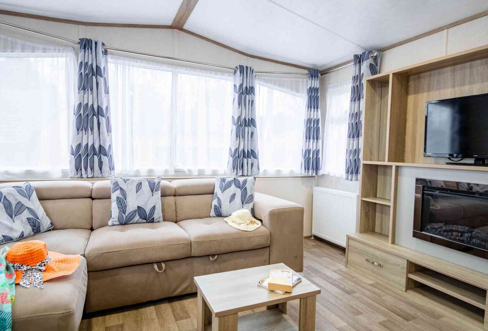 A cozy living area featuring a beige sofa with decorative cushions, a wooden coffee table, and a wall-mounted television. Large windows with sheer curtains allow natural light in, and the floor is covered with hardwood planks. Colorful items are scattered on the sofa and table.