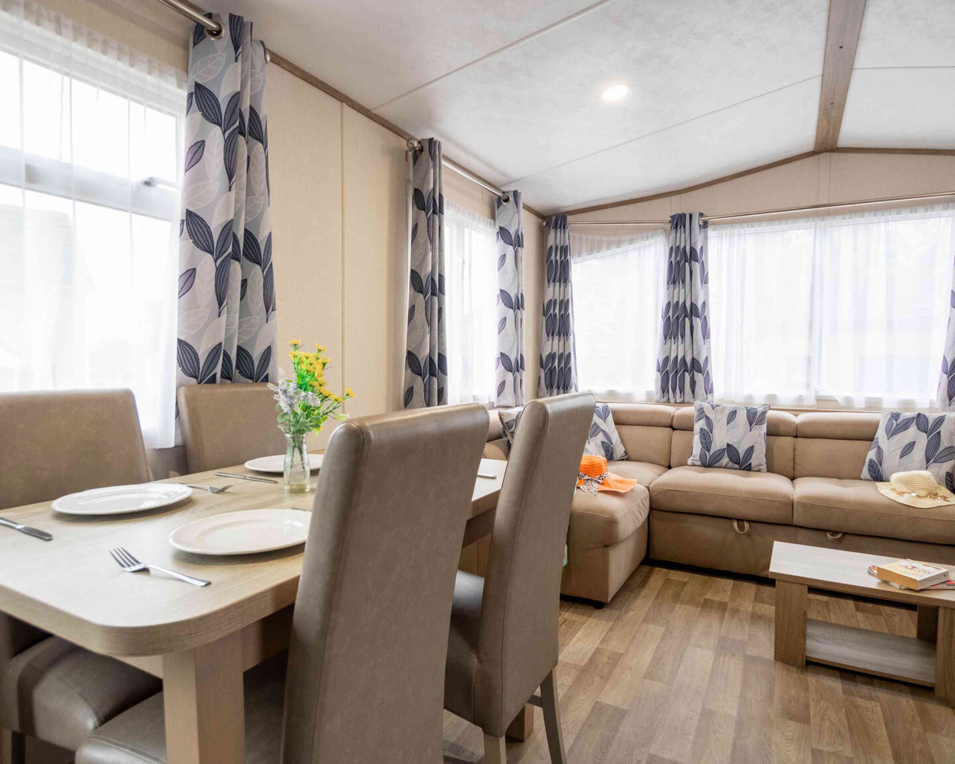 A cozy living area featuring a beige couch with patterned cushions, a wooden dining table set for several people, and large windows with sheer curtains adorned with leafy designs. A vase of flowers sits on the table, adding a touch of color.
