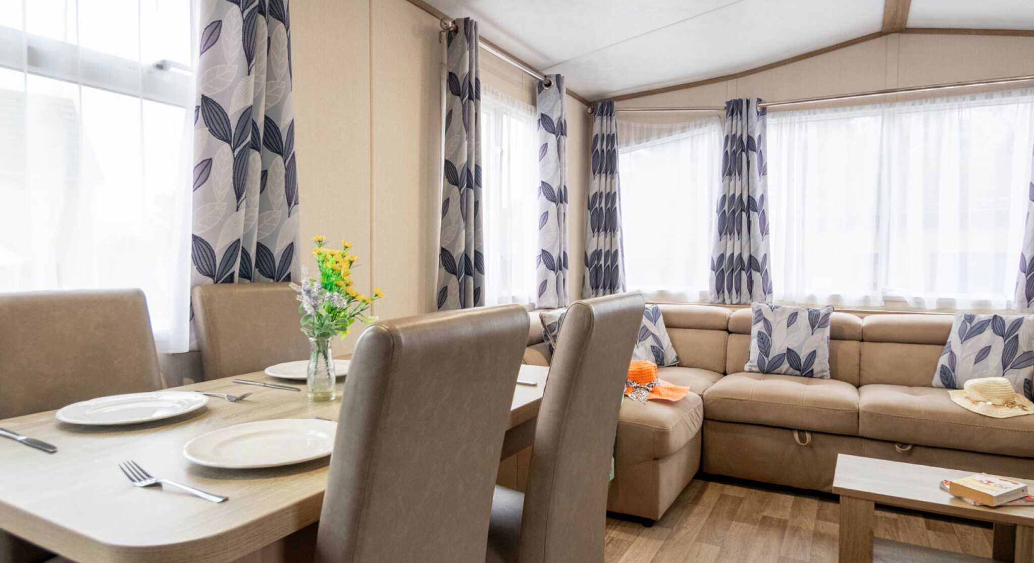 A cozy living area featuring a beige couch with patterned cushions, a wooden dining table set for several people, and large windows with sheer curtains adorned with leafy designs. A vase of flowers sits on the table, adding a touch of color.