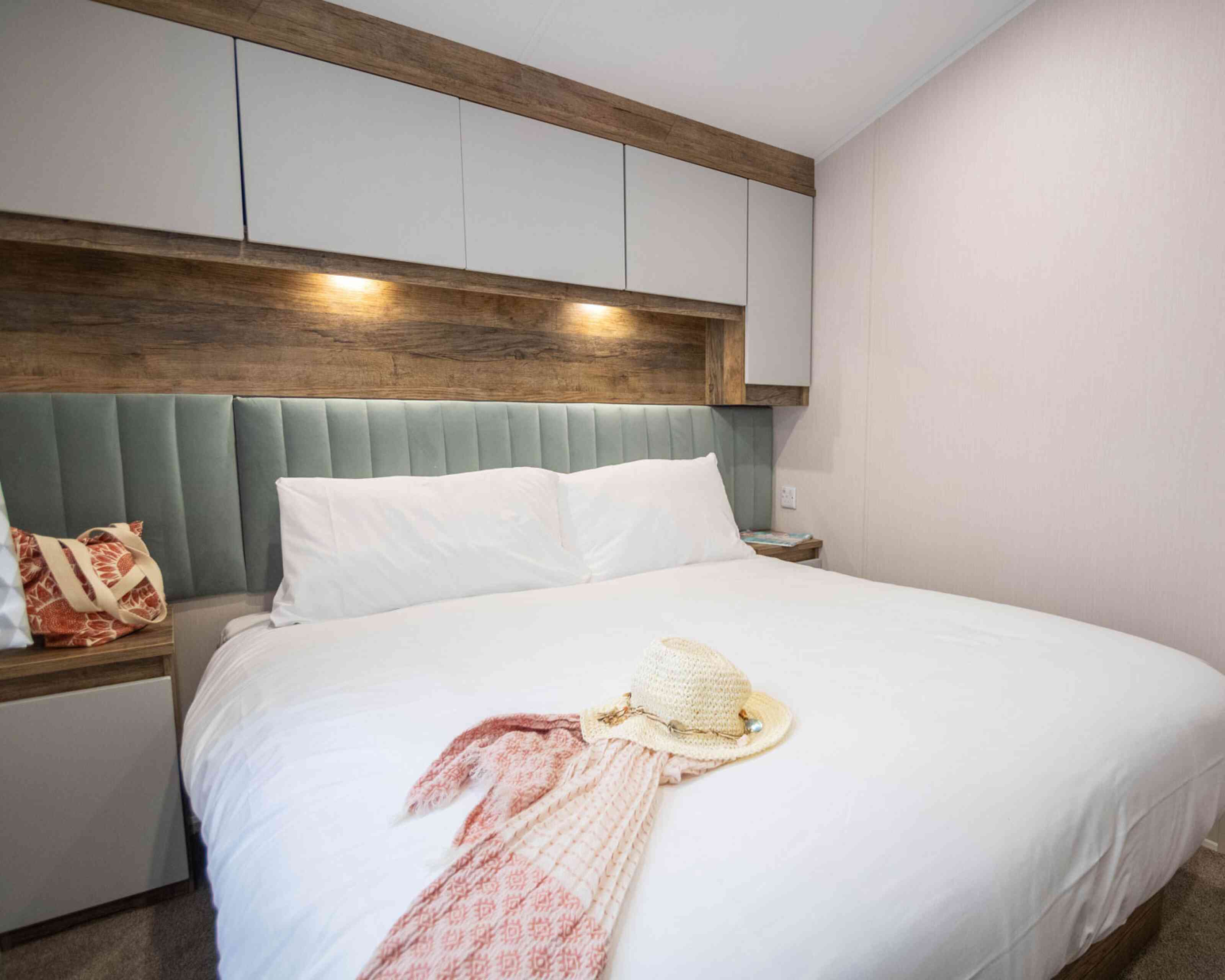 A cozy bedroom with a large bed featuring a light gray headboard and white bedding. A straw hat and a patterned scarf are placed on the bed. Soft lighting is installed above the headboard, and there's a wooden accent wall.