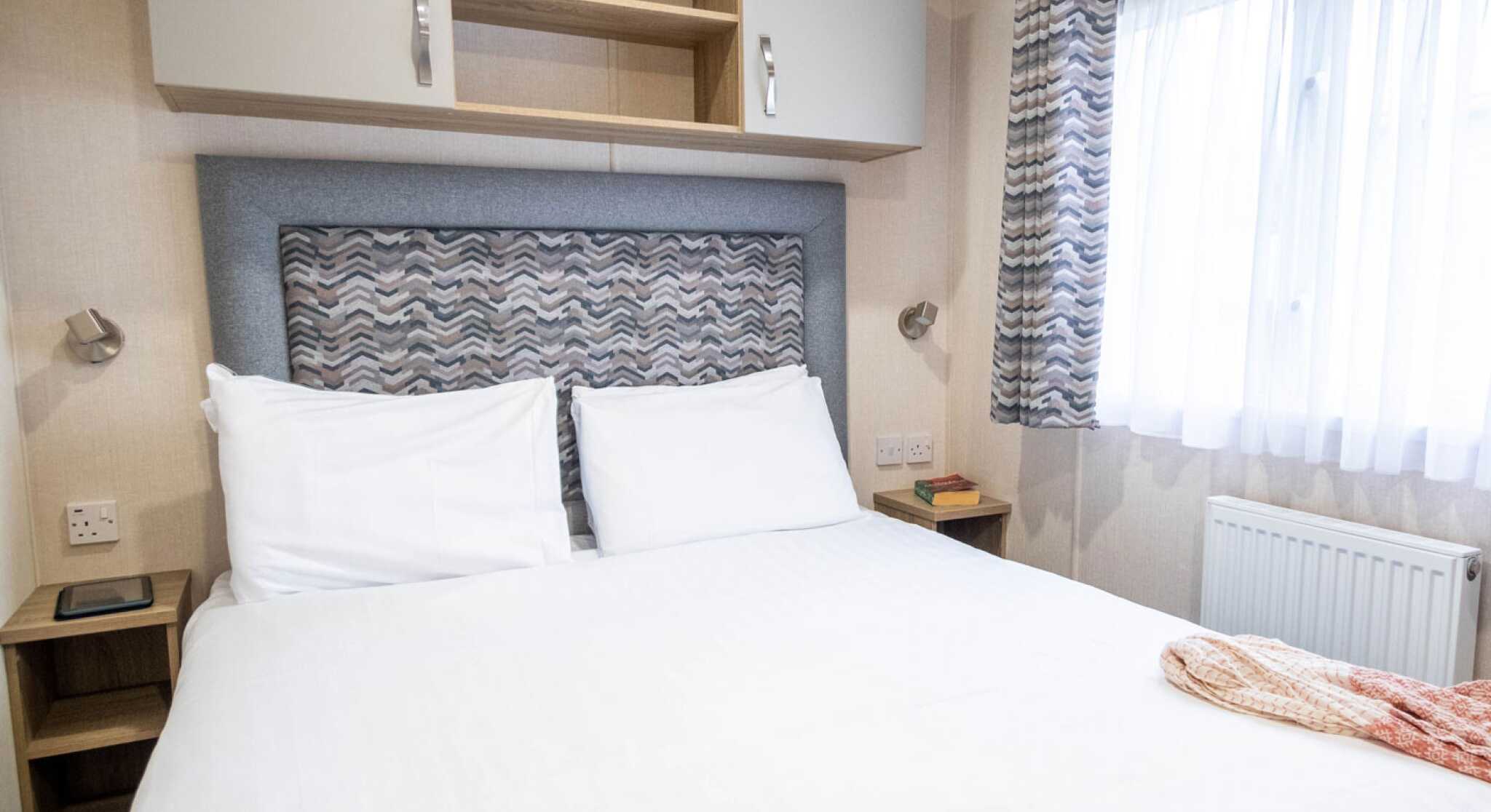 A neatly made double bed with white linens in a cozy room. Above the bed, there's a patterned headboard with light fixtures on either side. To the left, a small wooden nightstand holds a tablet. A window with sheer curtains allows natural light to enter, and wall-mounted cabinets are above the bed.