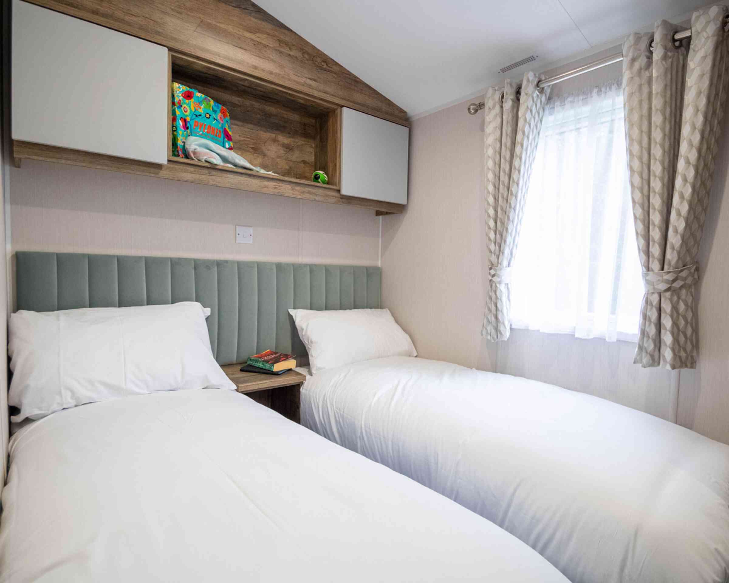 Two single beds with white bedding are positioned against a light-colored wall. Above the beds, a wooden shelf holds a colorful item and a small green decoration. A window with sheer curtains allows natural light to enter the room. A bedside table features a book and a decorative item.