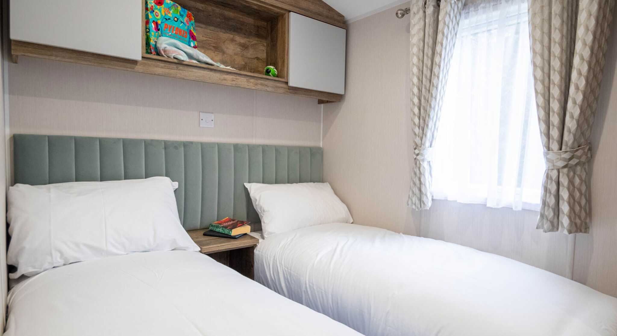 Two single beds with white bedding are positioned against a light-colored wall. Above the beds, a wooden shelf holds a colorful item and a small green decoration. A window with sheer curtains allows natural light to enter the room. A bedside table features a book and a decorative item.