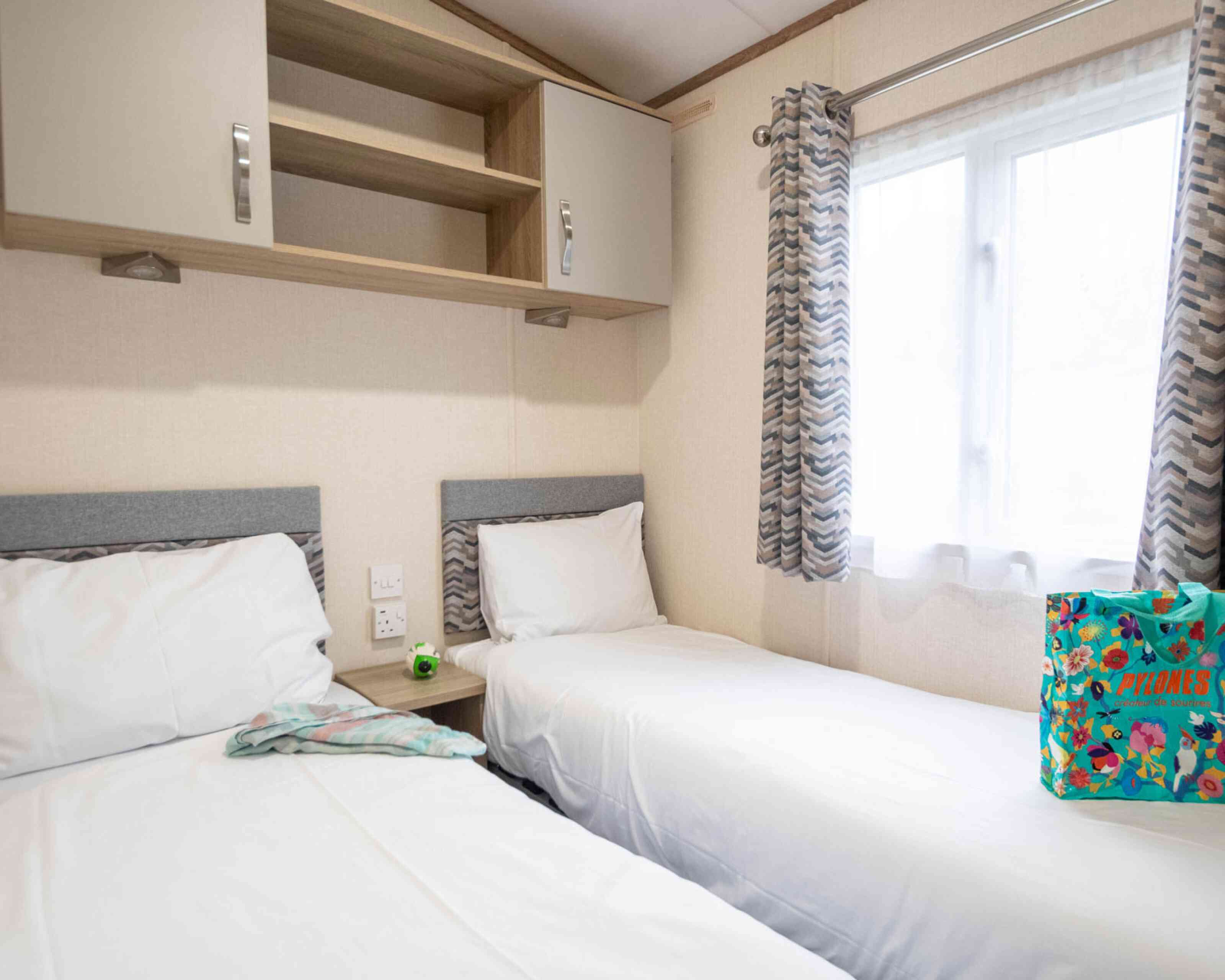 A cozy room featuring two single beds with white linens, a small bedside table, and overhead cabinets. A bright window with sheer curtains lets in natural light, and a colorful bag is placed on the floor next to one of the beds.