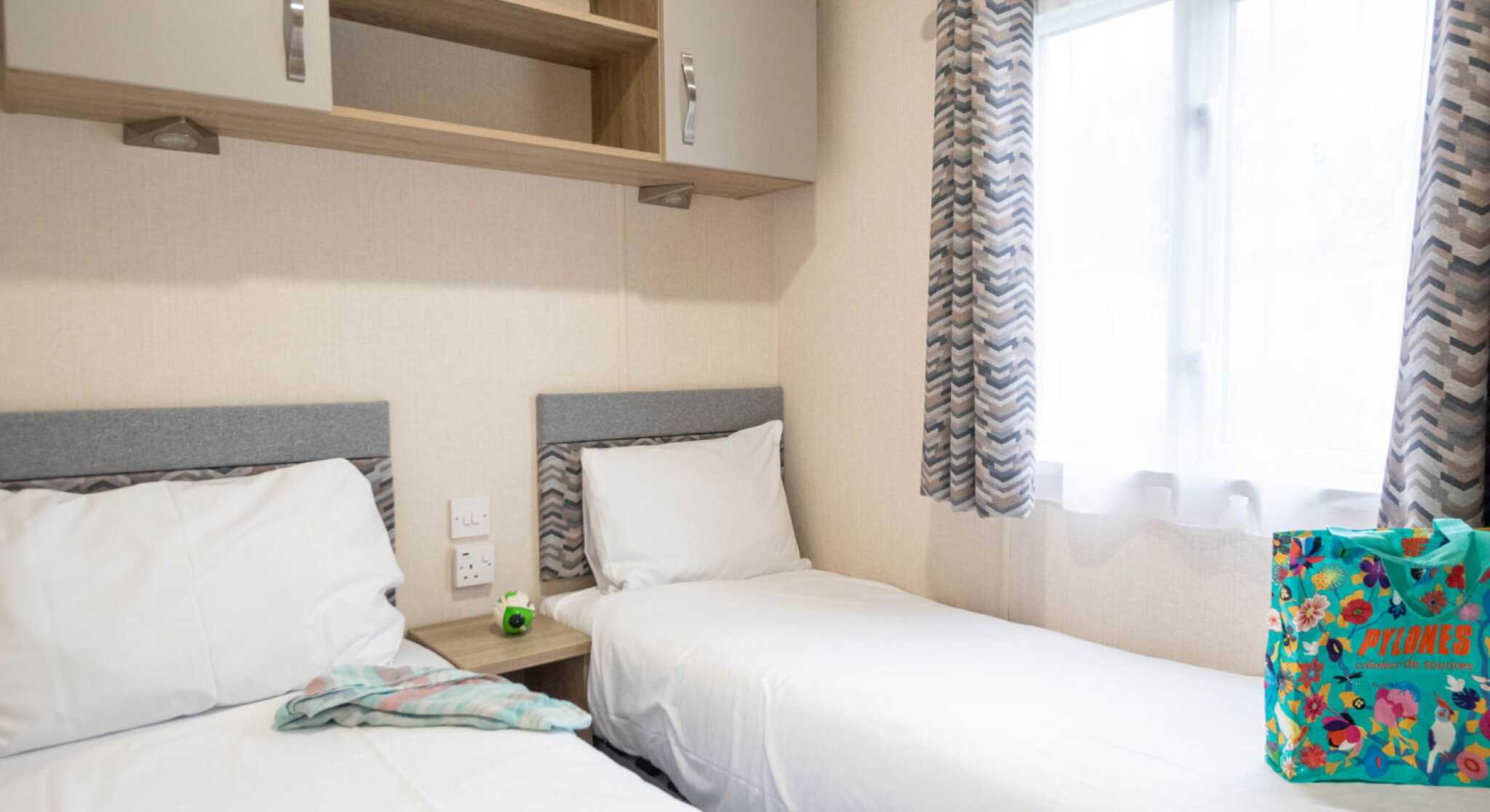 A cozy room featuring two single beds with white linens, a small bedside table, and overhead cabinets. A bright window with sheer curtains lets in natural light, and a colorful bag is placed on the floor next to one of the beds.