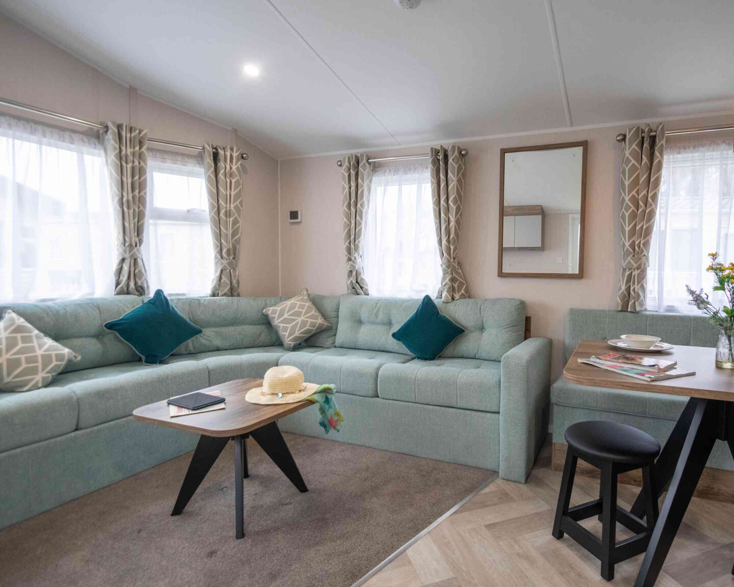 A cozy living space featuring a light green sectional sofa with decorative pillows, a wooden coffee table with a book and a hat, and a small dining area with a black stool. Natural light streams in through large windows fitted with sheer curtains. A mirror hangs on the wall beside the sofa.