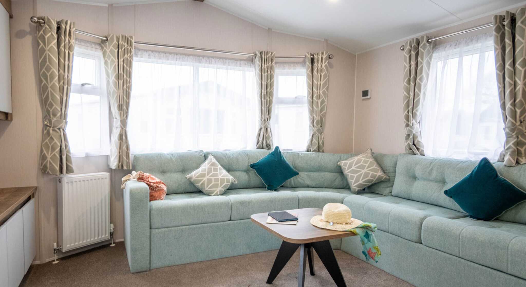 A cozy living room with a light blue sectional sofa adorned with decorative pillows. A round black coffee table sits in front, featuring a notebook and a sun hat. Large windows with sheer curtains allow natural light to fill the space.