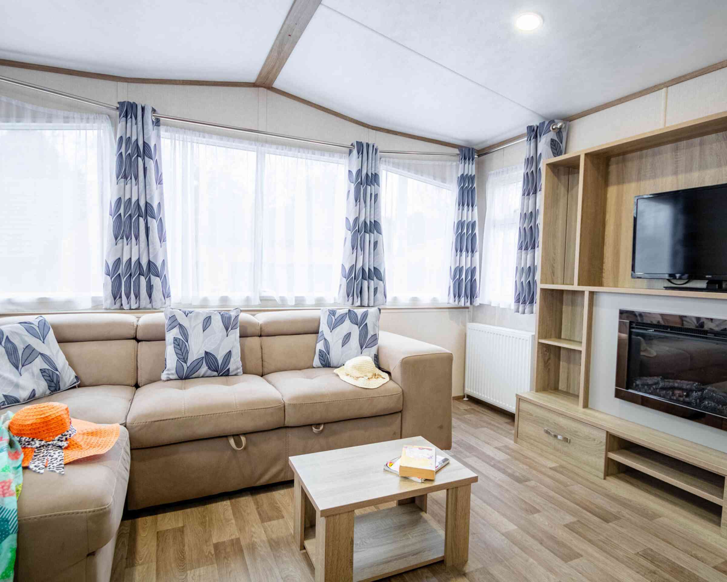 A cozy living room featuring a beige sectional sofa adorned with decorative cushions, a wooden coffee table, and a wall-mounted television. Sunlight streams through large windows covered with patterned curtains, while a bright, colorful throw and snacks add a homely touch.