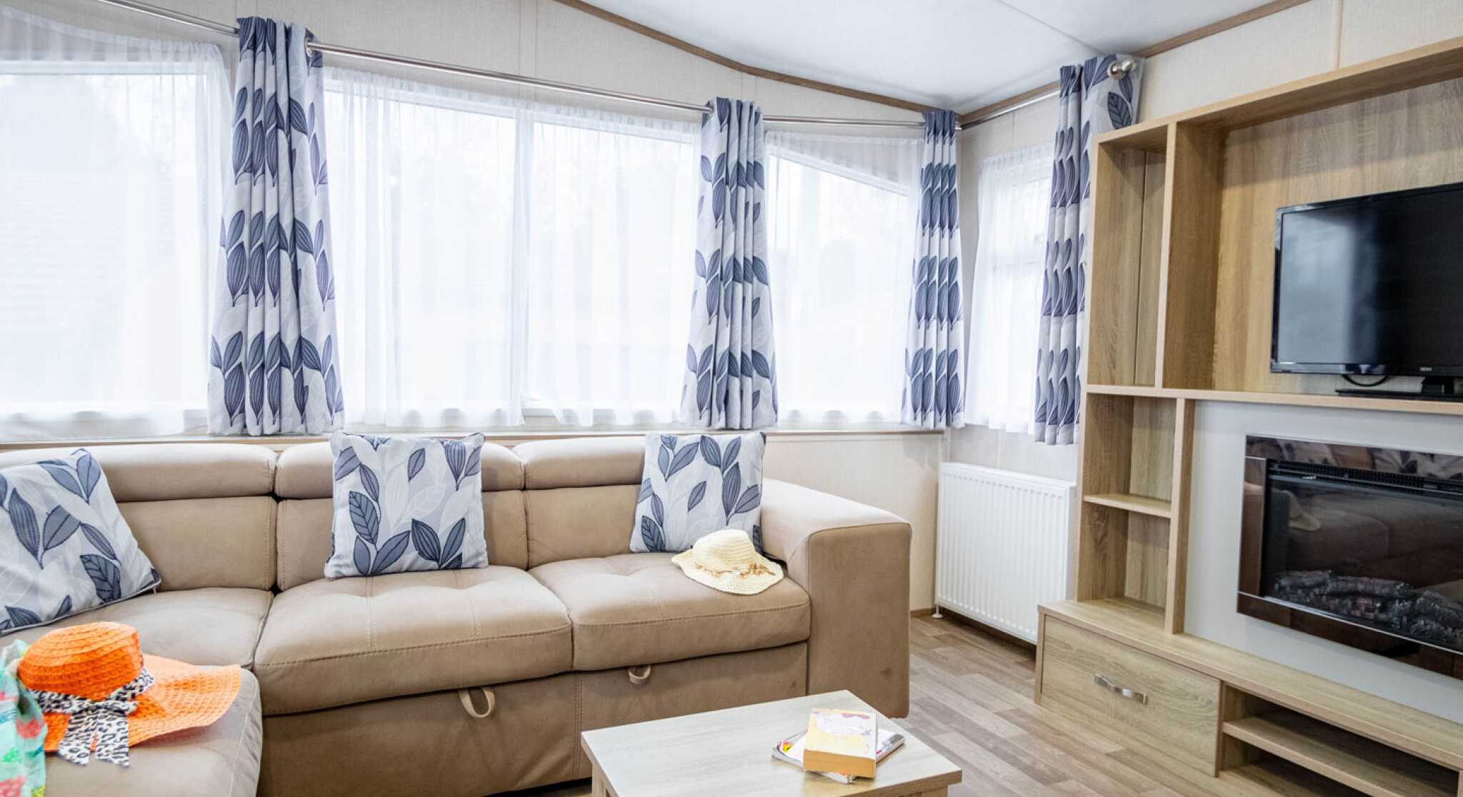 A cozy living room featuring a beige sectional sofa adorned with decorative cushions, a wooden coffee table, and a wall-mounted television. Sunlight streams through large windows covered with patterned curtains, while a bright, colorful throw and snacks add a homely touch.