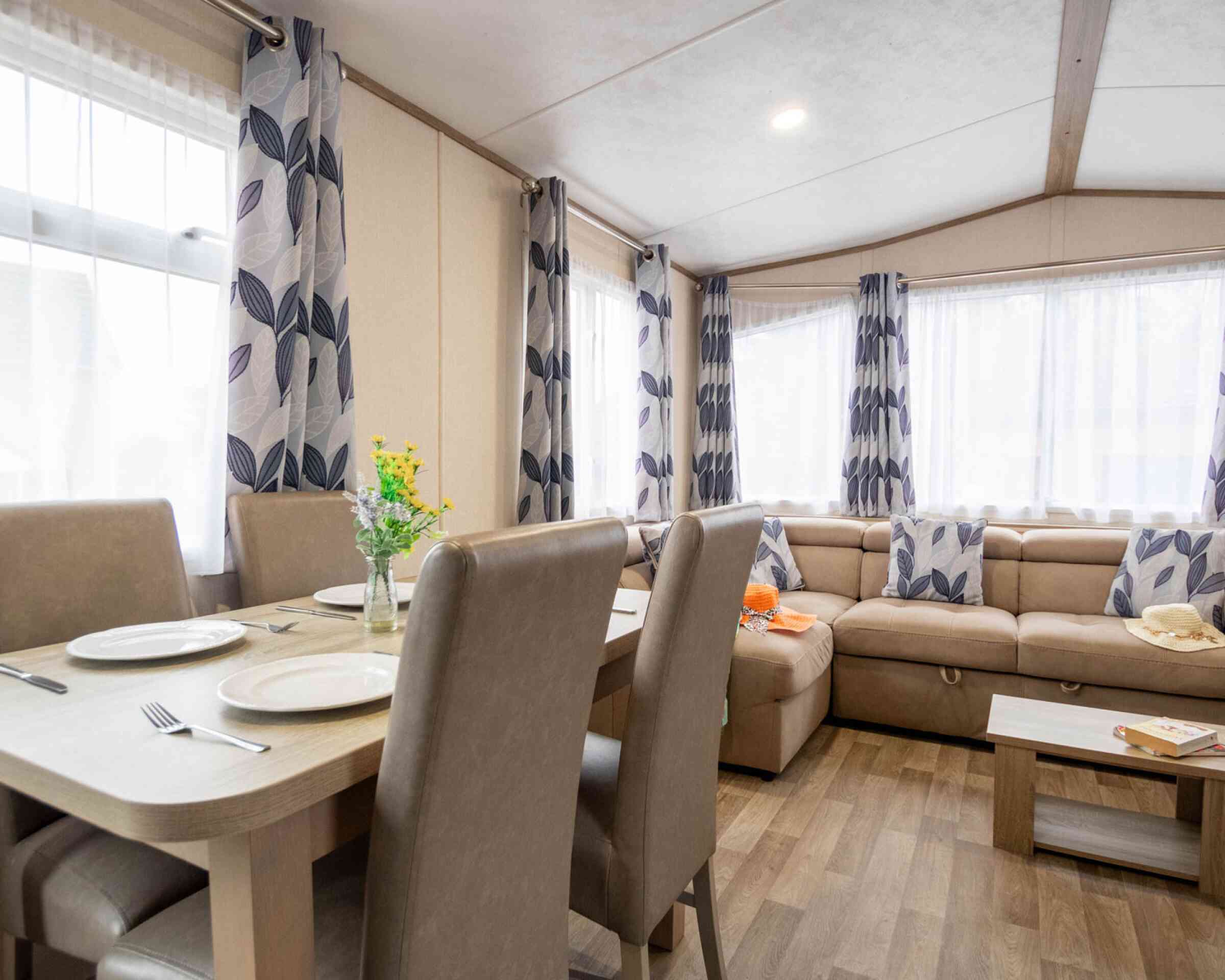 A cozy living area features a beige sofa adorned with patterned cushions, light curtains letting in natural light, and a dining table set for four. A floral centerpiece sits on the table, enhancing the inviting atmosphere.