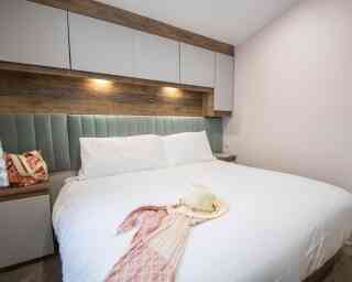 A neatly made bed with white linens and two plush pillows is featured in a cozy room. A straw hat and a patterned scarf are casually placed on the bed. The walls have a soft, neutral tone, accented by wooden panels and stylish lighting above the headboard.