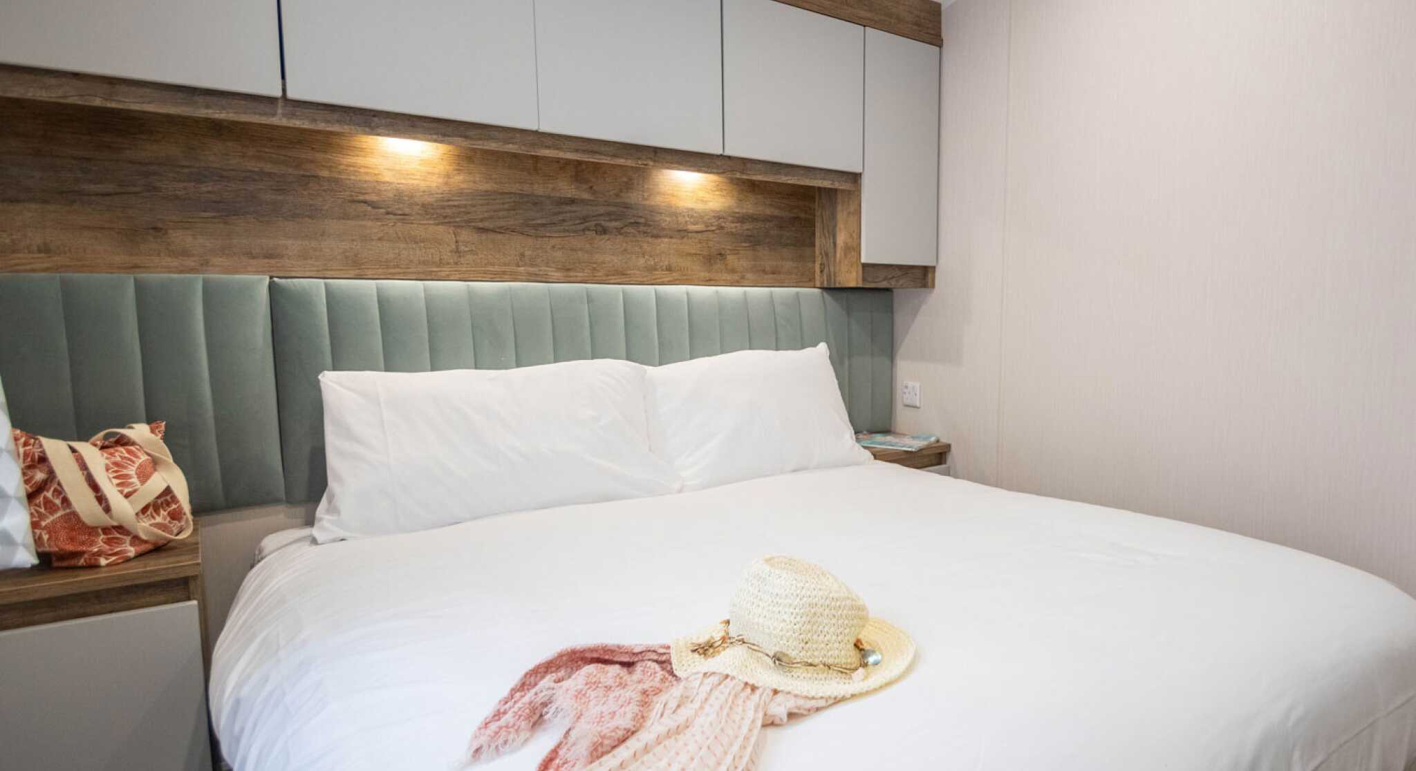 A neatly made bed with white linens and two plush pillows is featured in a cozy room. A straw hat and a patterned scarf are casually placed on the bed. The walls have a soft, neutral tone, accented by wooden panels and stylish lighting above the headboard.