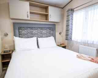 A neatly made double bed with white linens, featuring a patterned headboard, is positioned in a cozy room. There are bedside tables on either side and warm-toned curtains covering a window. Shelves above the bed contain storage, while a soft throw is draped over the bed.