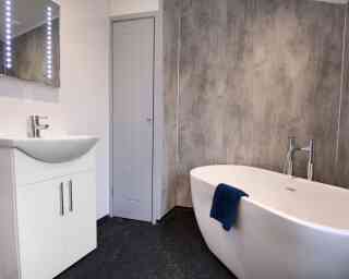 A modern bathroom featuring a freestanding white bathtub, a sleek vanity with a sink, and a mirror with backlit lights. The walls have a textured gray finish, and a blue towel drapes over the bathtub. A gray door is visible in the background.