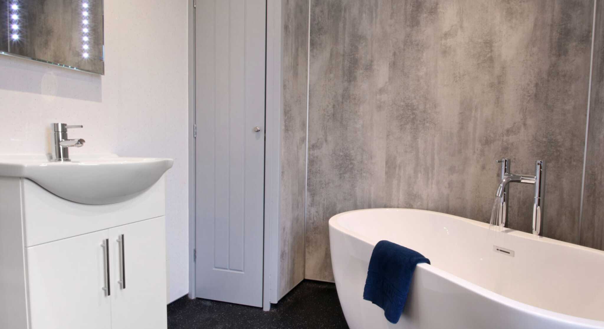 A modern bathroom featuring a freestanding white bathtub, a sleek vanity with a sink, and a mirror with backlit lights. The walls have a textured gray finish, and a blue towel drapes over the bathtub. A gray door is visible in the background.