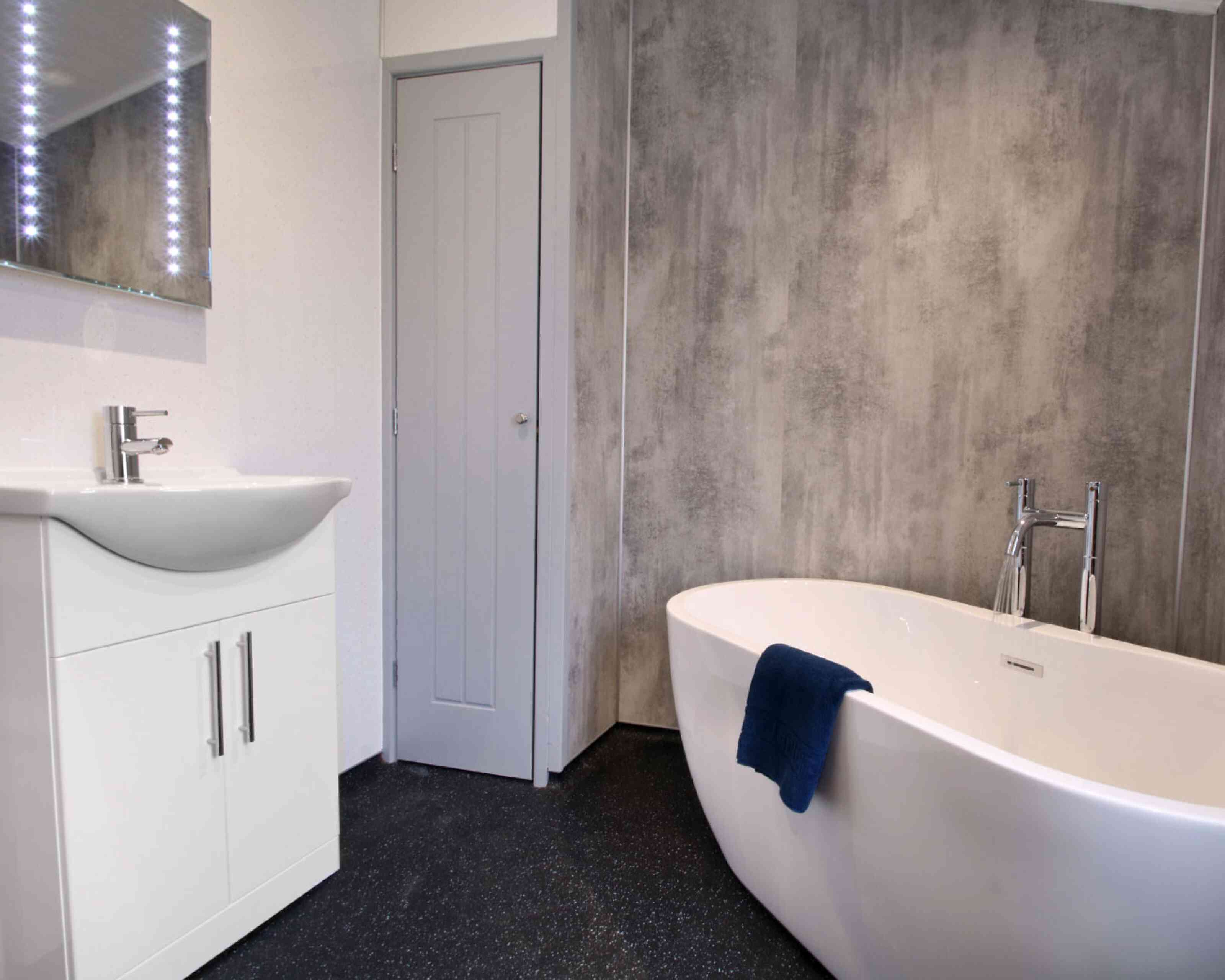A modern bathroom featuring a freestanding white bathtub, a sleek vanity with a sink, and a mirror with backlit lights. The walls have a textured gray finish, and a blue towel drapes over the bathtub. A gray door is visible in the background.