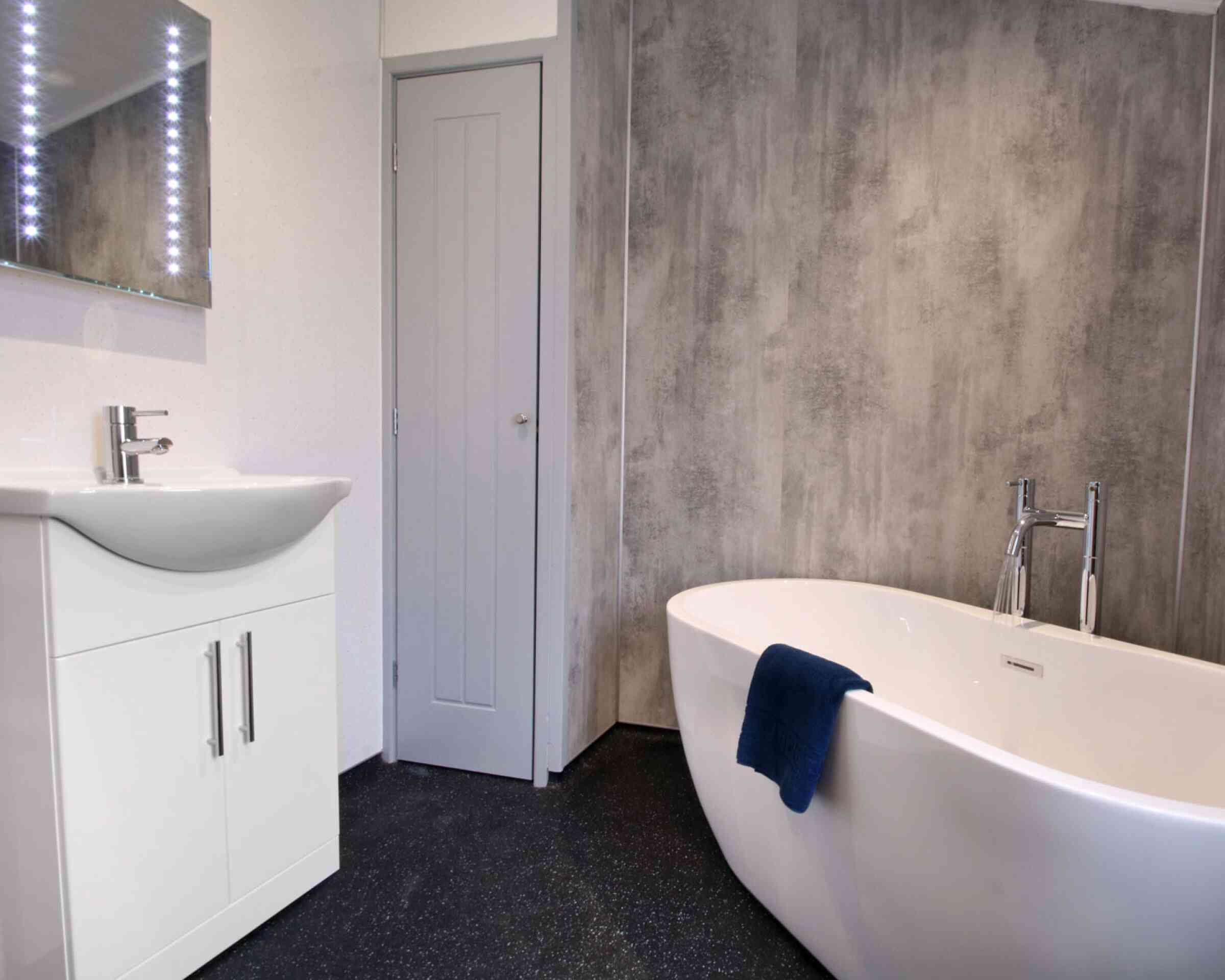 A modern bathroom featuring a freestanding white bathtub, a sleek vanity with a sink, and a mirror with backlit lights. The walls have a textured gray finish, and a blue towel drapes over the bathtub. A gray door is visible in the background.