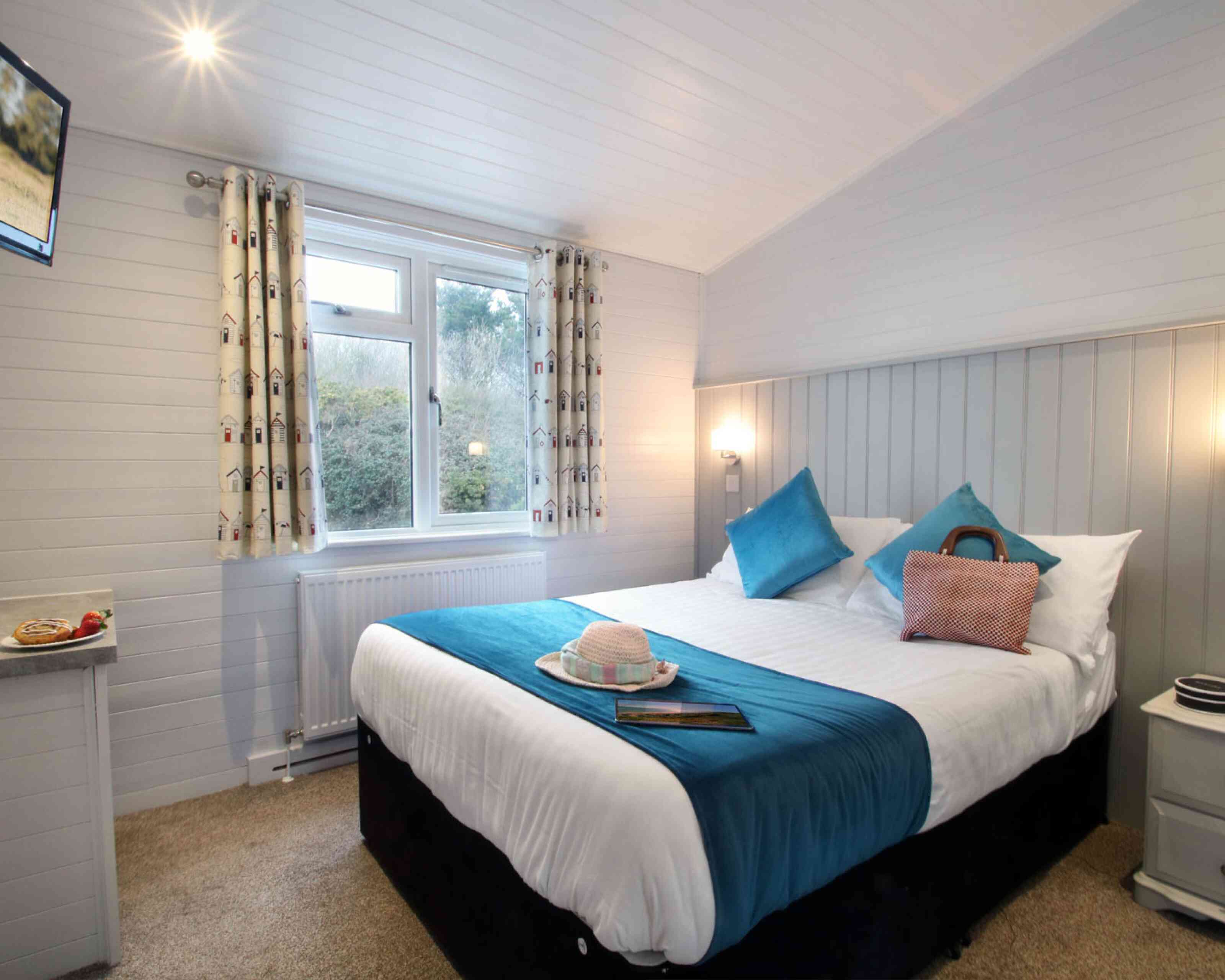 A cozy guest room featuring a double bed with white linens and turquoise accents, complemented by decorative pillows. There is a small table next to the bed with a book and a sun hat, and a window dressed with patterned curtains overlooks greenery outside. A flat-screen TV is mounted on the wall.